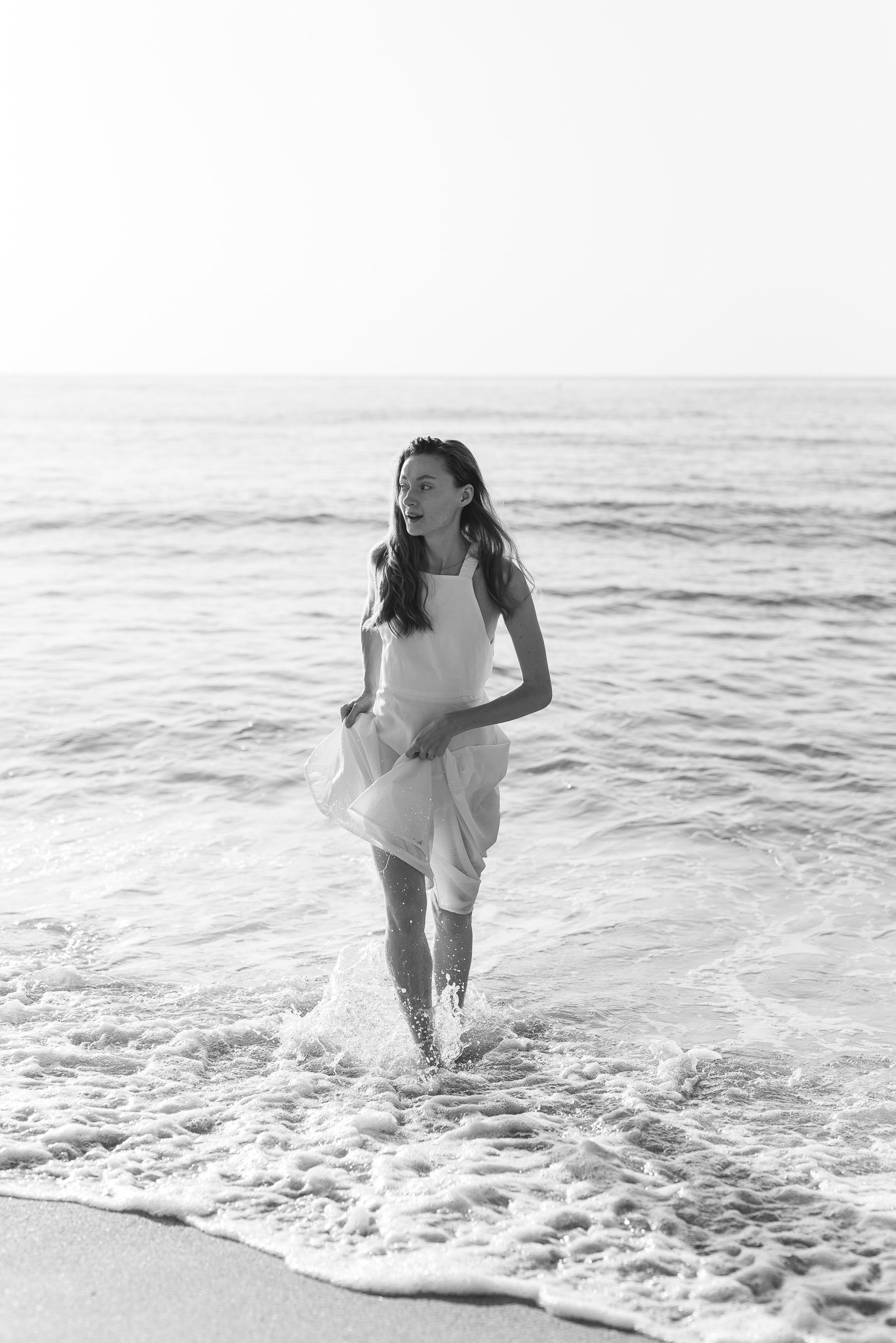 A woman in a white dress walking out of the ocean on a beach, splashing water with her feet.