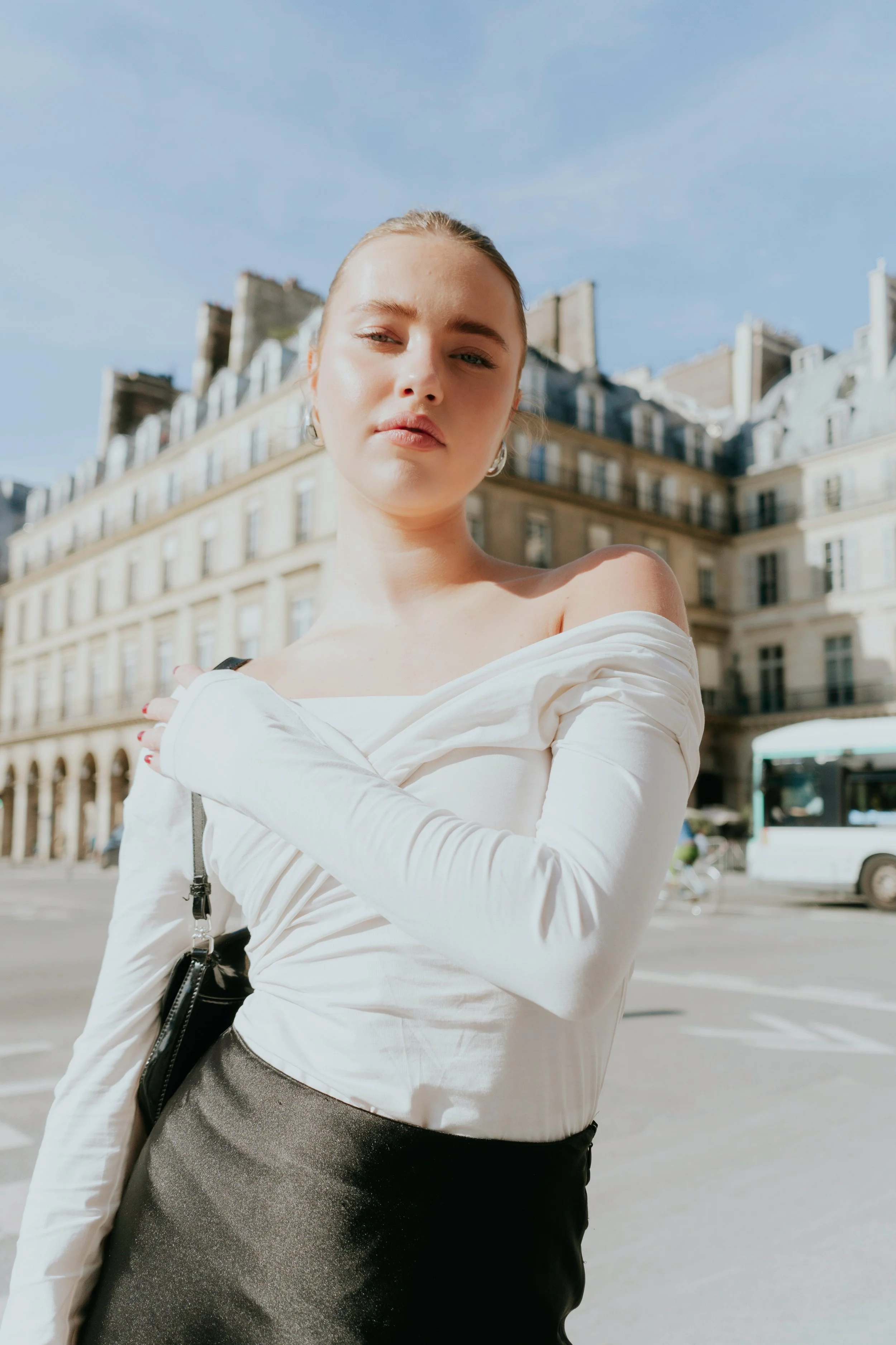 A young woman with blonde hair and earrings standing outside on a city street with historic European-style buildings in the background.