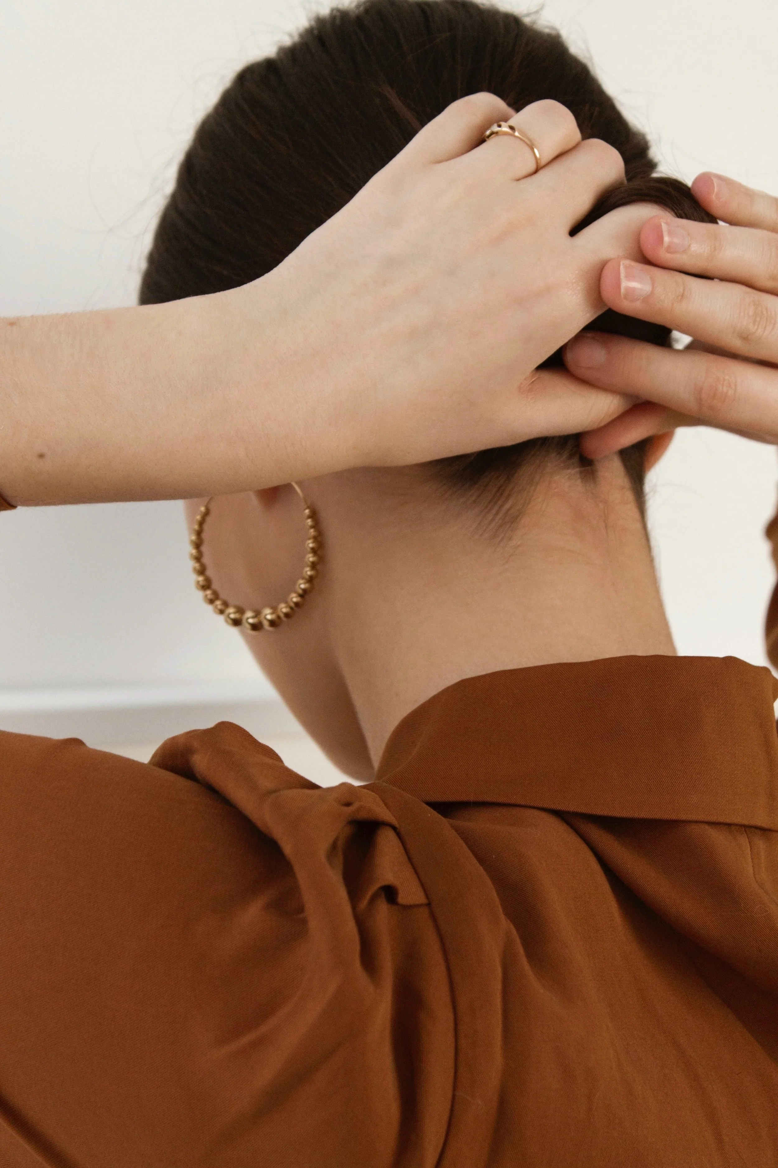 Person touching the back of their head and neck, wearing earrings and a gold ring, dressed in a brown shirt.
