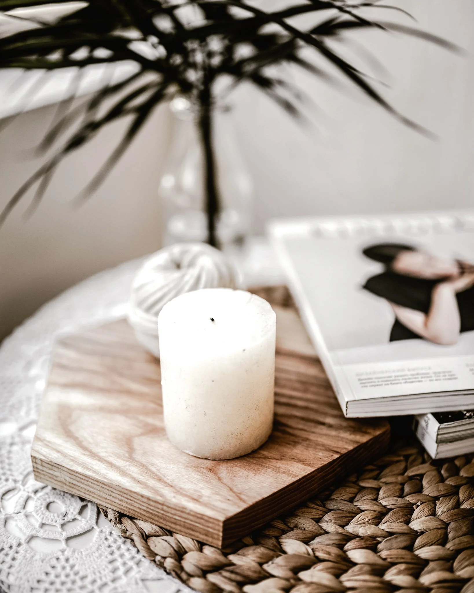 Decorative arrangement with a white candle on a wooden board, a plant with long leaves, and a magazine on a woven placemat.