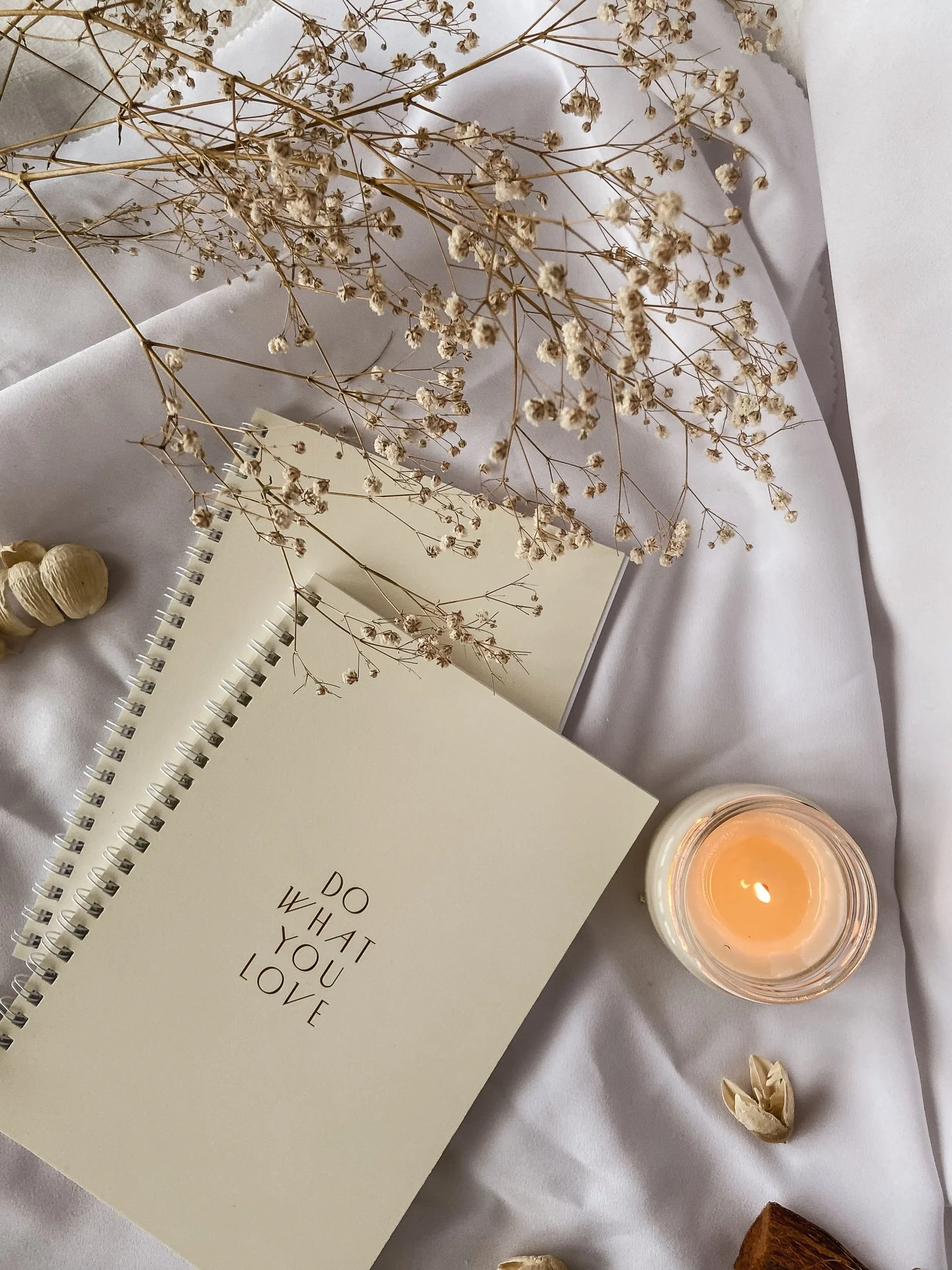Minimalist flat lay with a white notebook titled "Do What You Love," dried flowers, a lit candle, and small dried decorative items on white fabric.