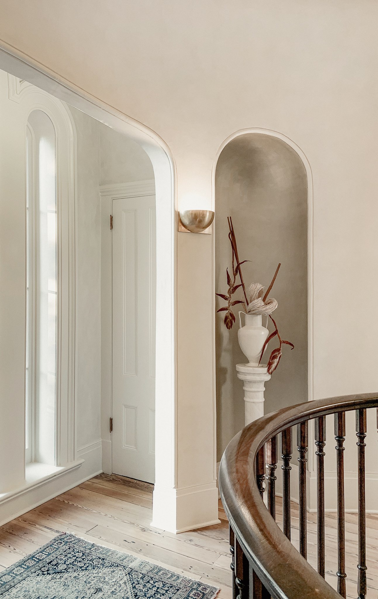 Interior view of a staircase landing with a white vase holding dried decorative plants on a pedestal, an arched wall opening, a wall sconce, and part of a wooden staircase railing.