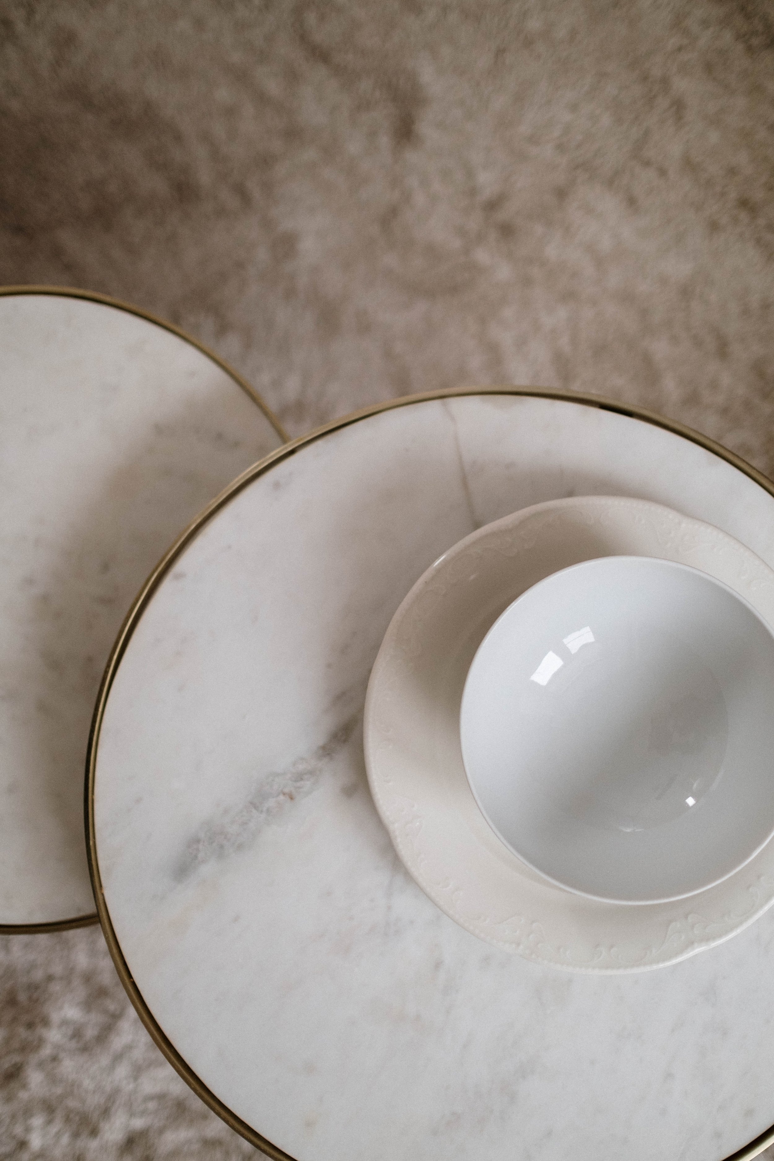 Close-up of a set of nested white plates with a decorative rim, placed on a small marble table with gold trim, on a beige carpeted floor.