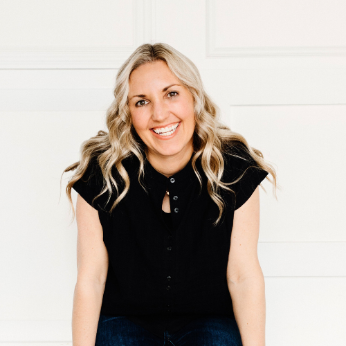 A smiling woman with long wavy blonde hair wearing a black shirt, sitting against a white background.