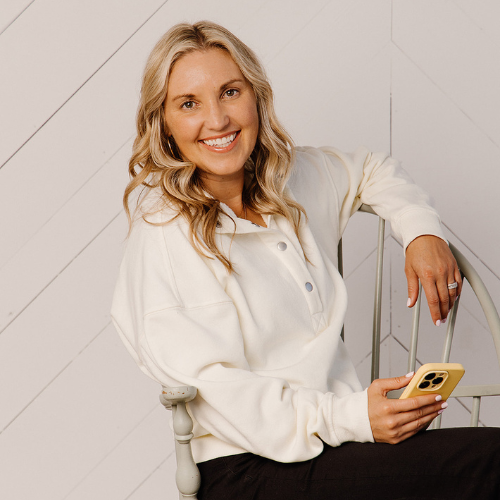 A smiling young woman with blonde hair in a white sweatshirt, sitting on a chair, holding a yellow smartphone, against a white wooden wall background.