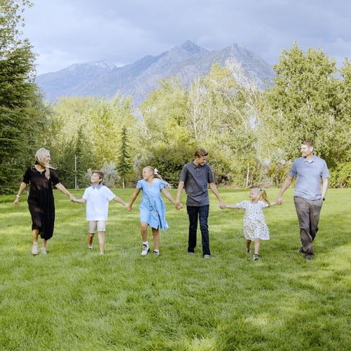 Family holding hands and walking together on a grassy field with trees and mountains in the background.