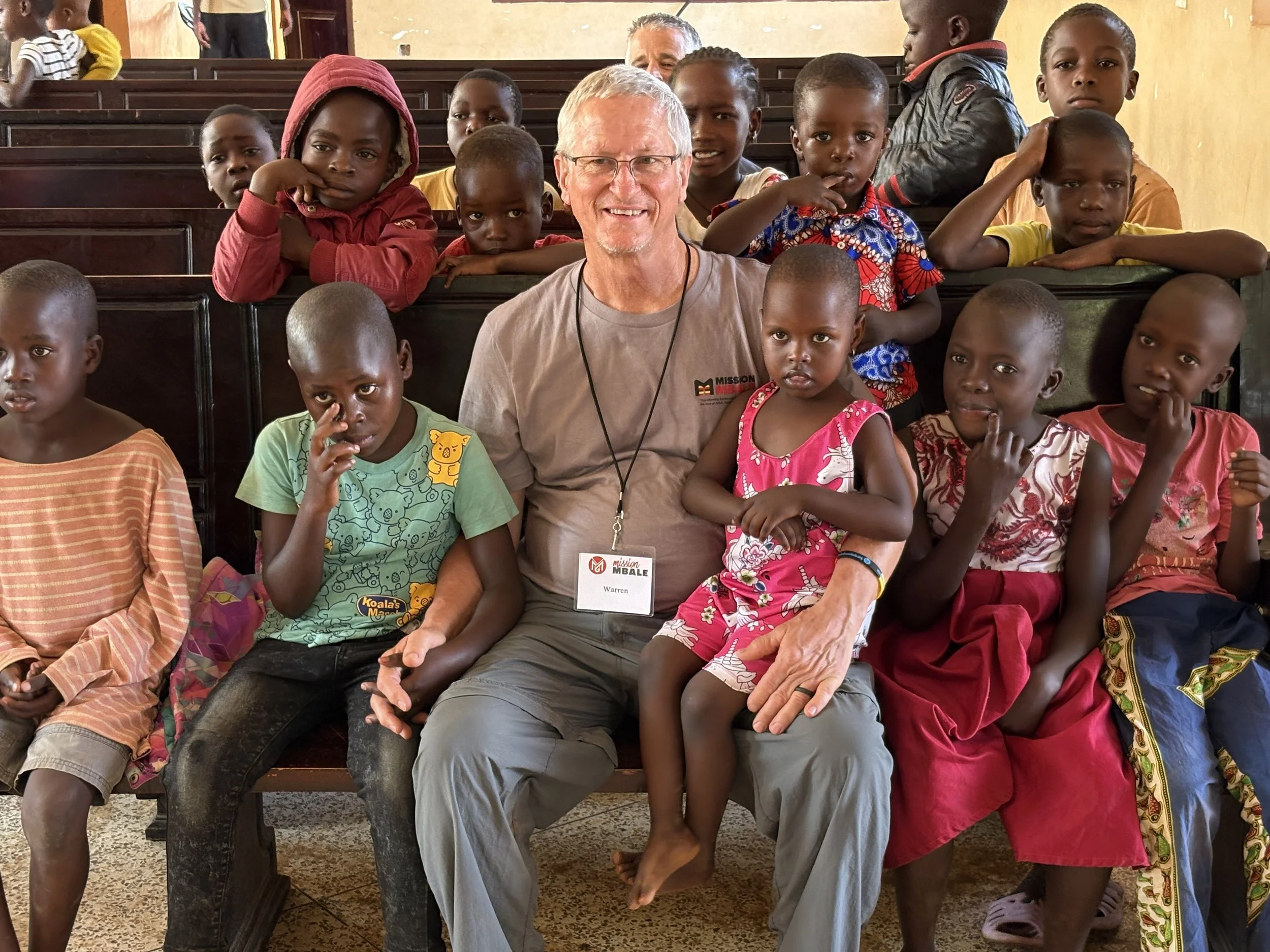 A man with glasses and gray hair, wearing a gray T-shirt and a name badge, is sitting in a church pew surrounded by children. The children, of African descent and various ages, are sitting and standing around him, some looking at the camera and others looking away. The setting appears to be a church with wooden pews and a beige wall.