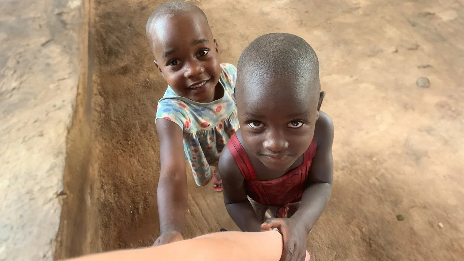 Two young children with shaved heads, one girl wearing a colorful dress and one boy wearing a red outfit, standing on a dirt floor inside a room, holding hands and looking up at the camera.