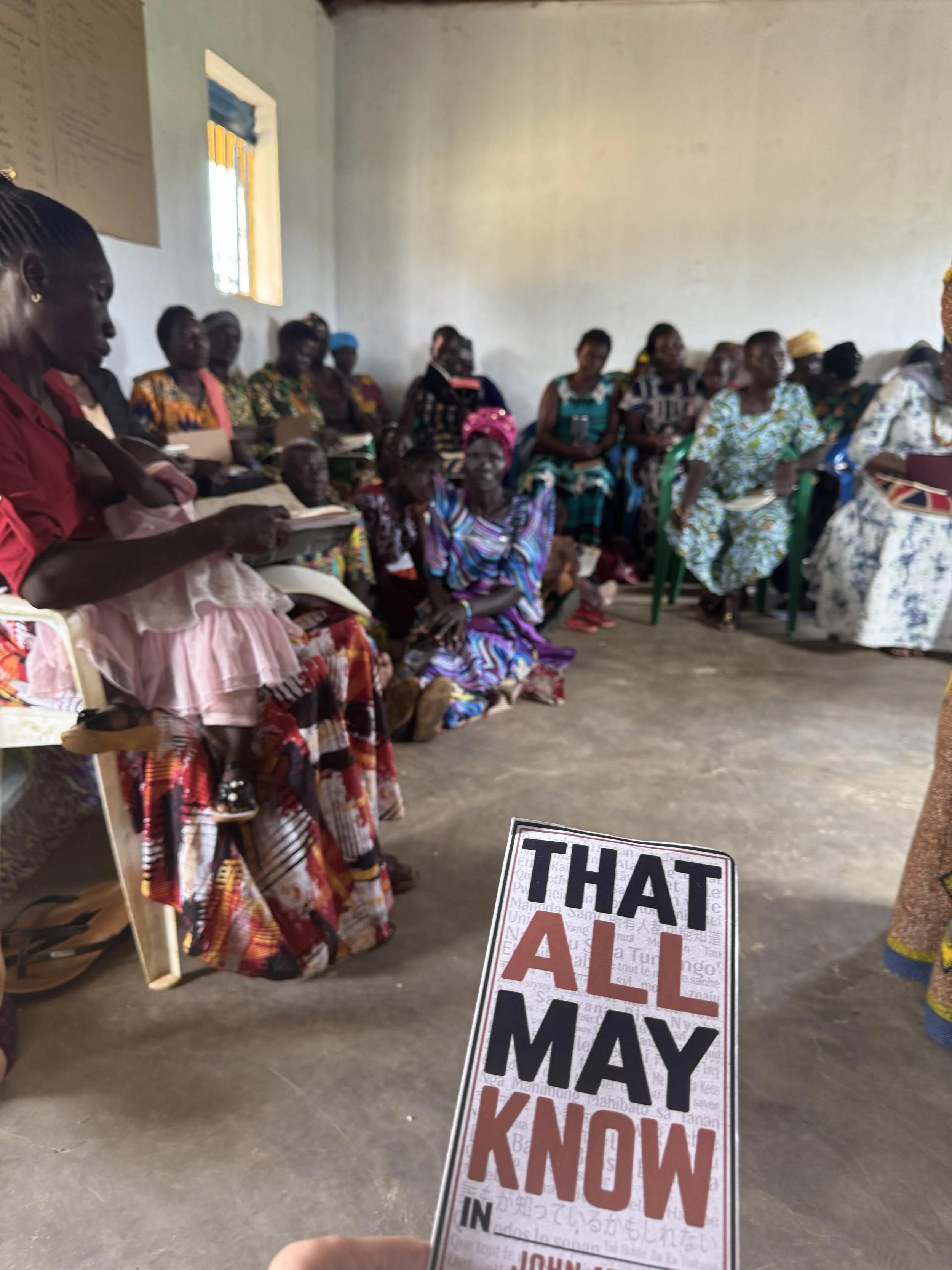 A group of women in colorful dresses seated and standing in a room, attending a gathering or meeting. A hand holding a card reads "That All May Know in" in the foreground.