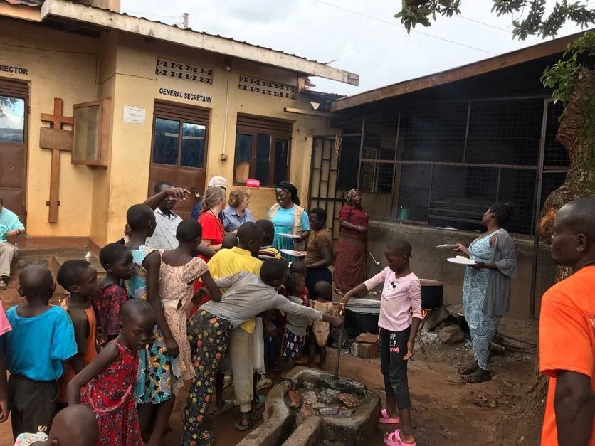 Group of children and adults gathered outdoors near a building with religious symbols. Children are fetching water from a communal well, while adults observe or assist. Building has signs indicating it is a church office.