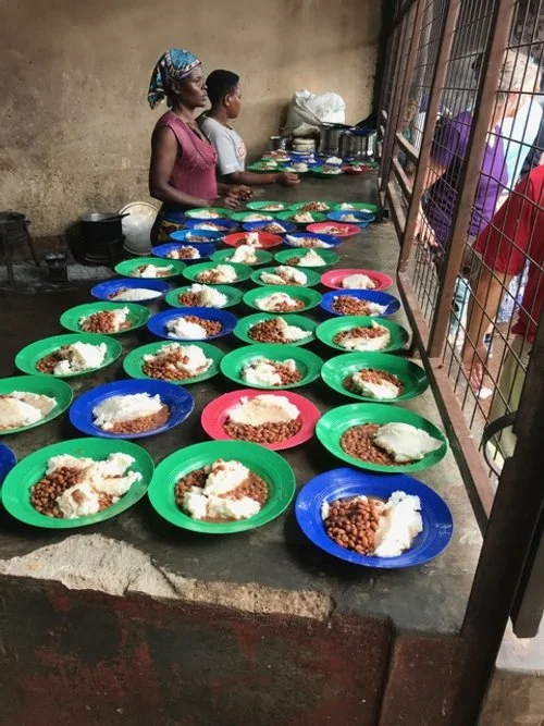 Women and children standing behind a table with multiple colorful plates of food, inside a building with a concrete wall and metal bars on the window.