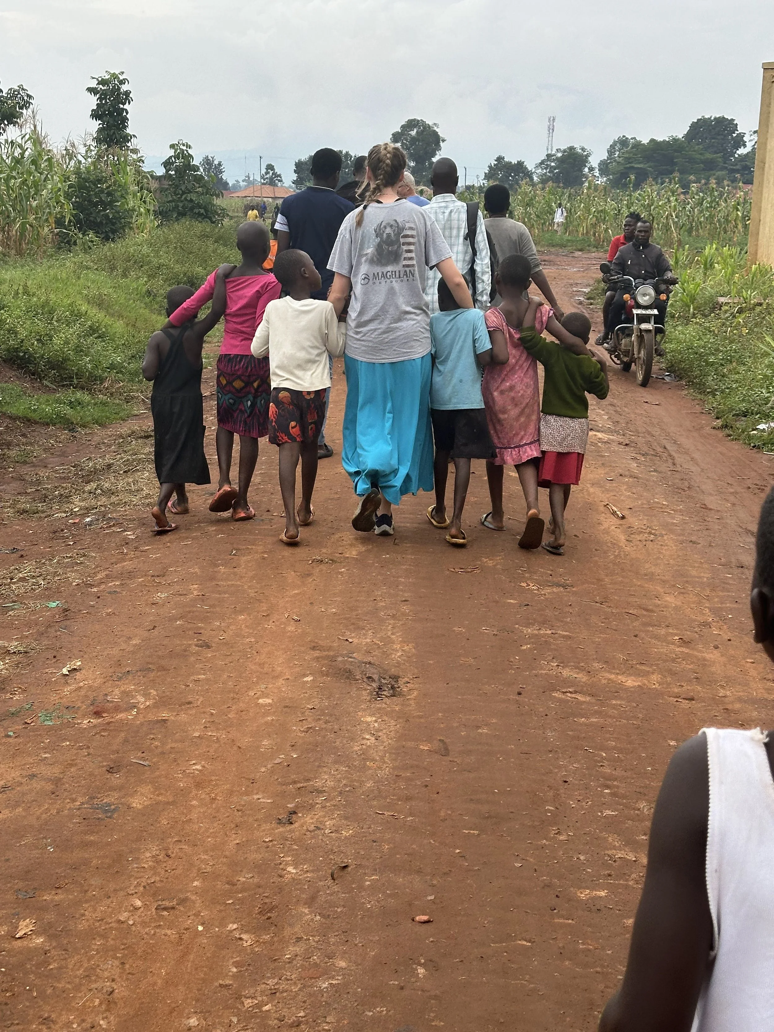 A group of children and adults walking down a dirt road in a rural area, with some children holding hands with the adults.