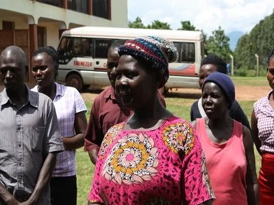 A group of people standing outdoors in front of a bus, with a woman in a pink floral shirt and a colorful head covering in the center.