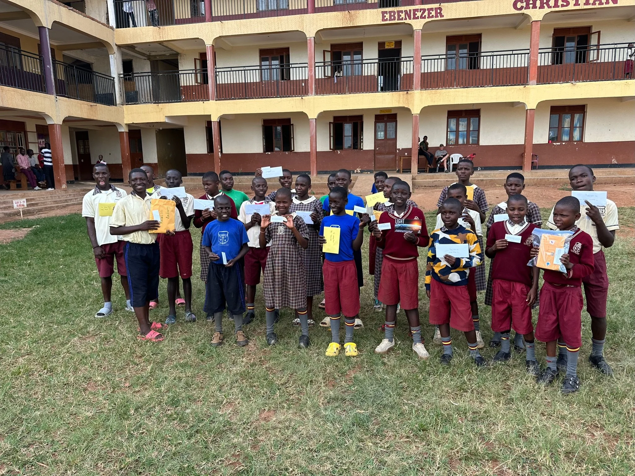 Group of school children standing outdoors on grass in front of a school building. The children are holding papers and smiling. The school building has balconies and the name 'Ebenizer Christian' visible on the top.