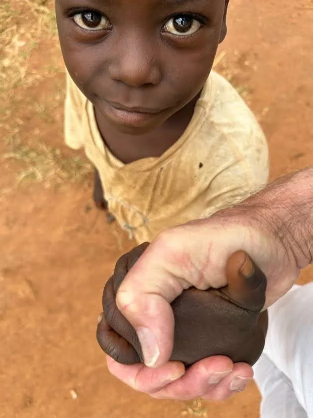A young boy with large expressive eyes looking up at the camera while an adult's hand holds his small dark-skinned hand.
