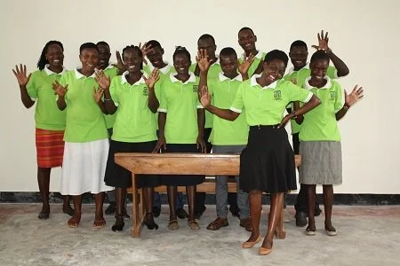 Group of women and girls wearing green shirts, standing together indoors, some smiling and waving, with a wooden table in front of them.