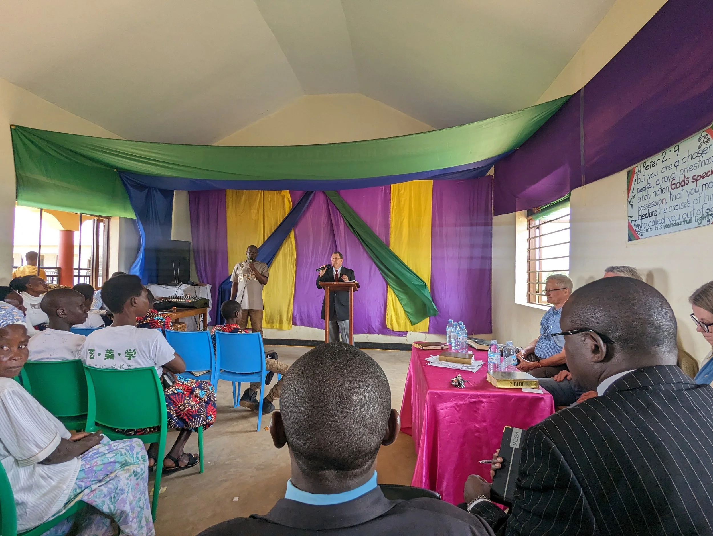 A gathering inside a church or community hall with a speaker at a wooden podium, people seated, some holding books and water bottles, with colorful drapes and a banner on the wall.
