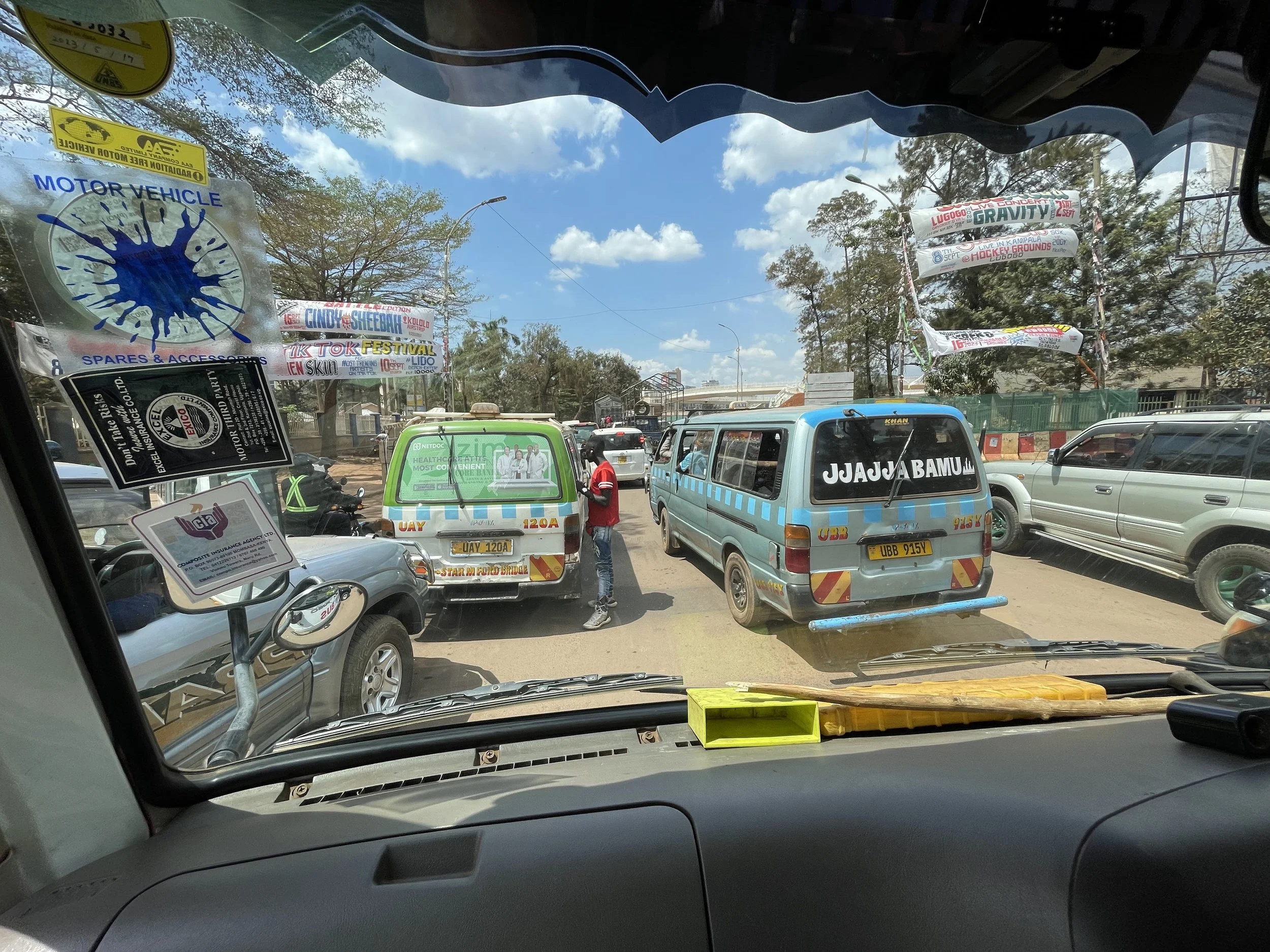 Street view from inside a vehicle, showing taxis and cars on a busy road with colorful banners and trees in the background.