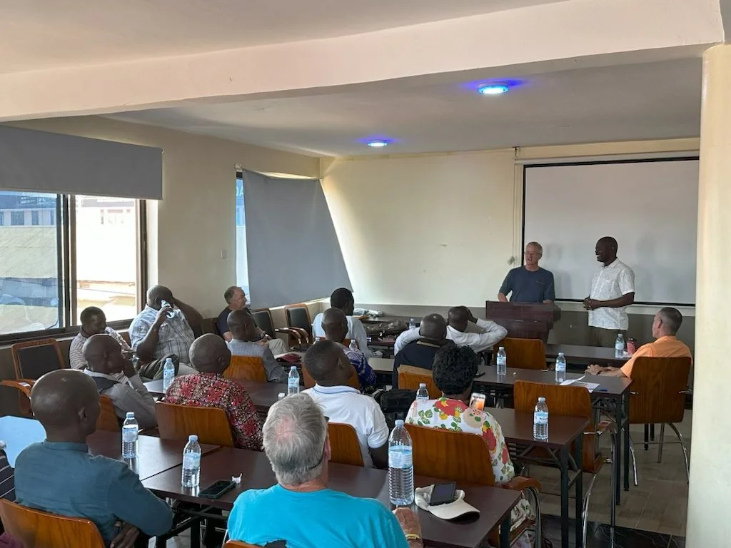 A group of people attending a presentation or lecture in a conference room, with two men standing at a podium in front of a white projection screen.