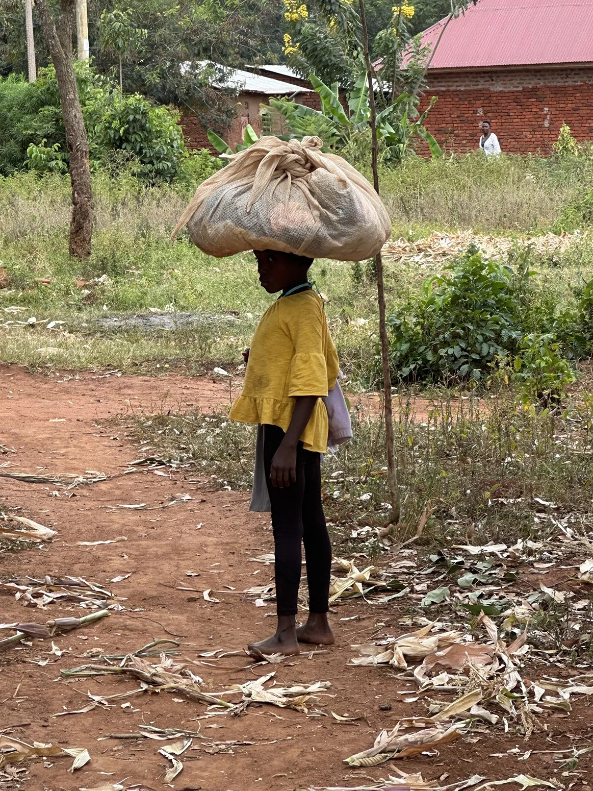 A young girl balancing a large bag on her head while standing barefoot on a dirt path in a rural area with trees, plants, and a brick building in the background.