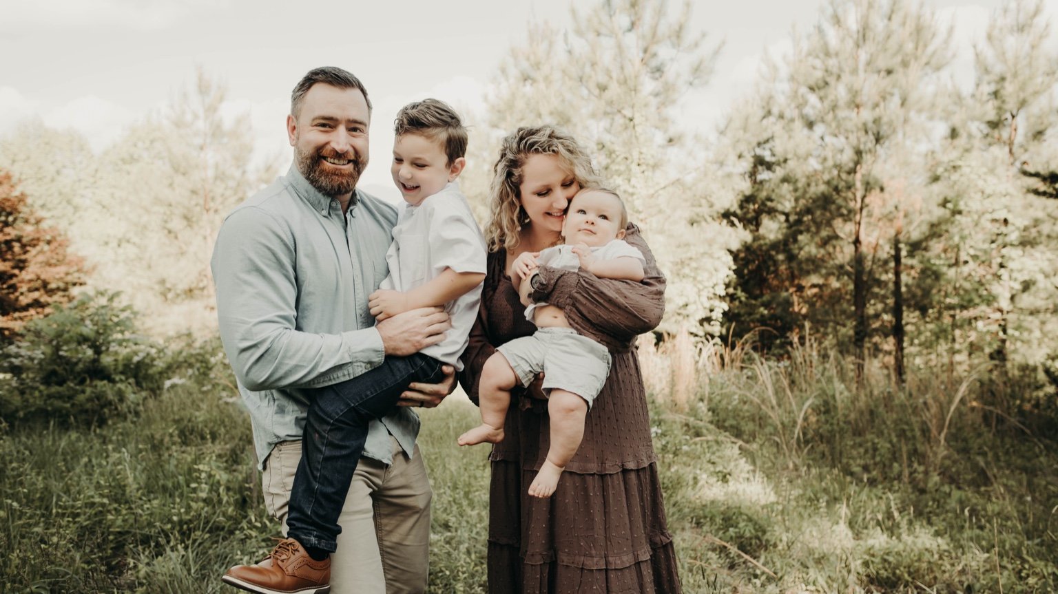 A happy family of four standing outdoors among trees and grass, with the father holding a young boy and the mother holding a baby, all smiling and enjoying a sunny day.