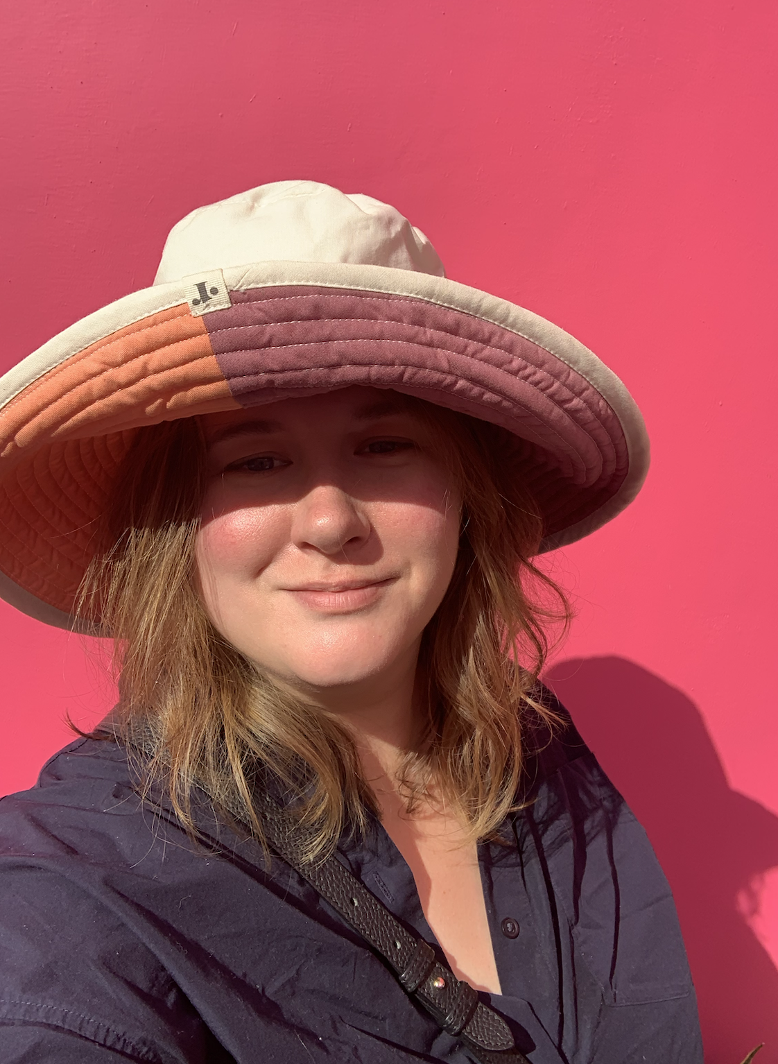 A woman with wavy red hair wearing a large wide-brimmed hat with sections of pastel orange, purple, and cream, standing against a bright pink wall.