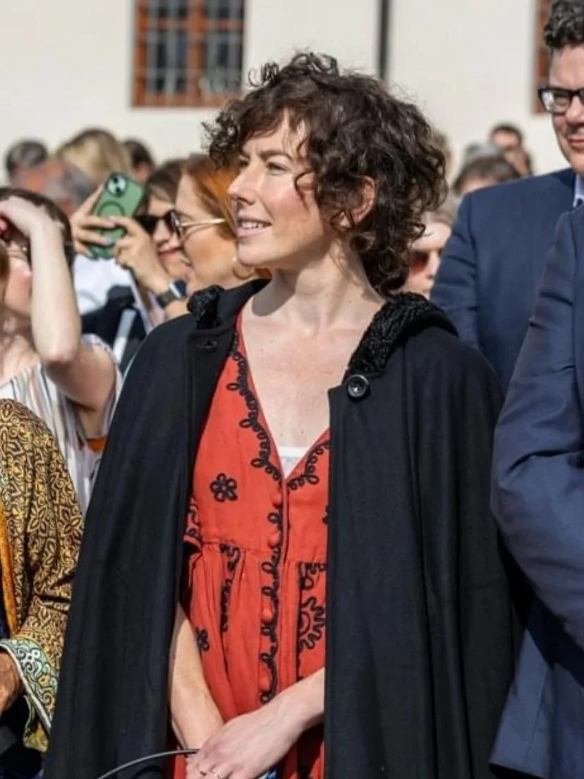 A woman with curly hair wearing a red dress with black embroidery, standing outdoors at an event, with people around her, some taking photos.