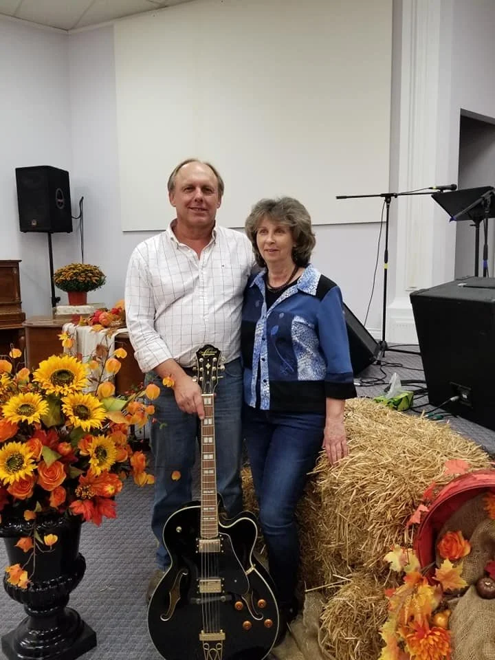A man and woman standing indoors with a guitar, fall-themed decorations, including flowers, a hay bale, and pumpkins, and a microphone and speaker setup in the background.