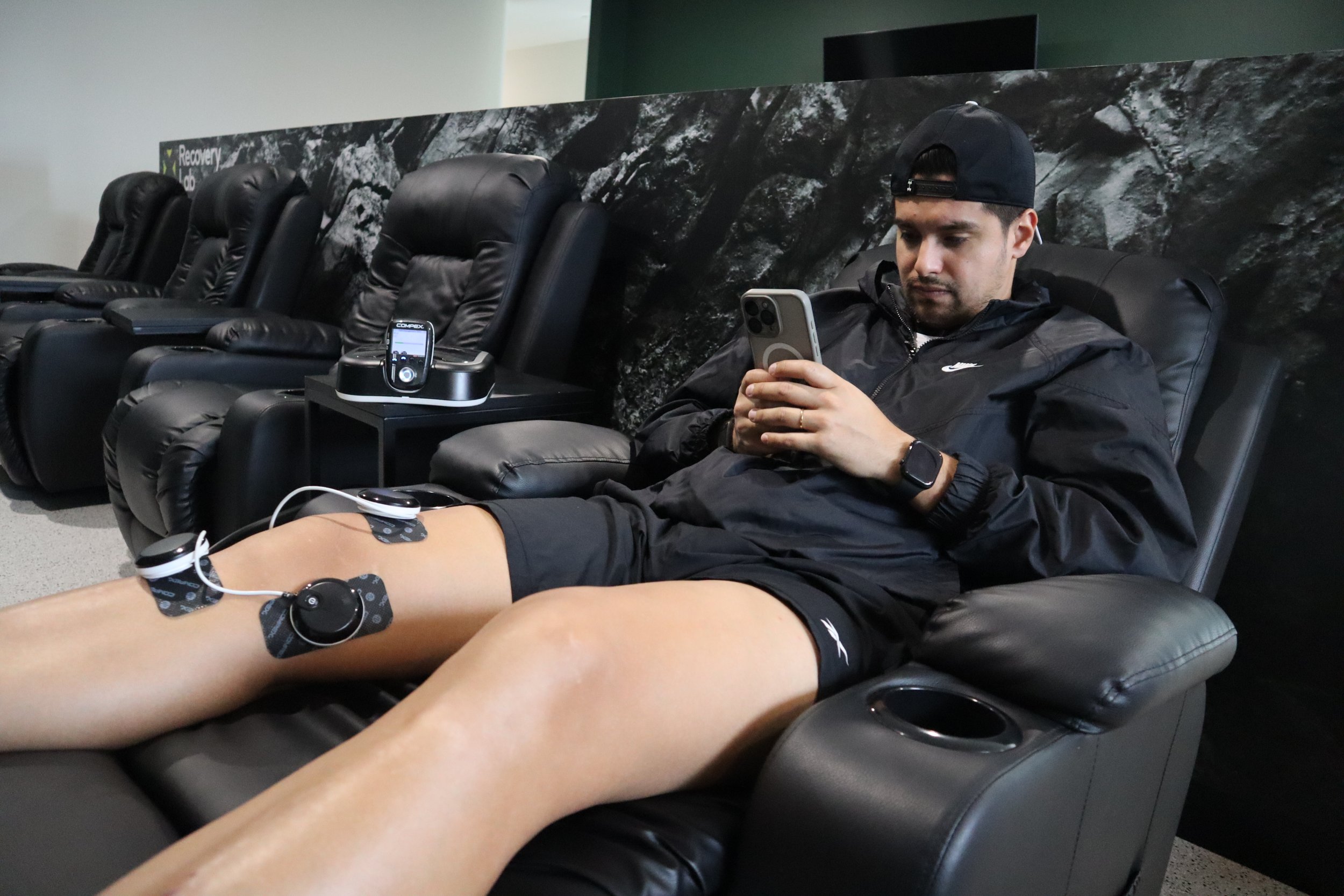 A man sitting in a black recliner at a recovery lounge, using his smartphone with his legs elevated and connected to a medical device with electrodes and wires.