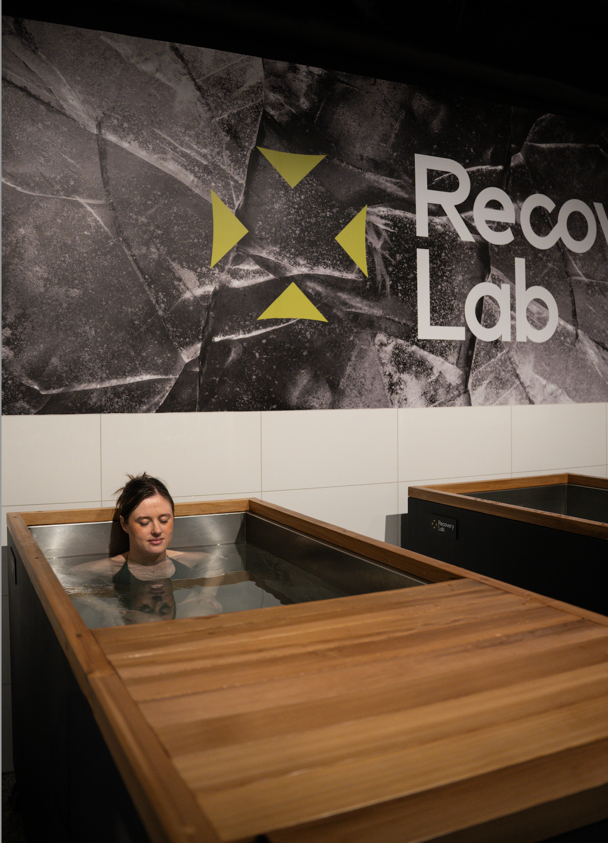 Woman relaxing in a wooden hot tub at a recovery lab, with a large sign on the wall that reads 'Recovery Lab'.