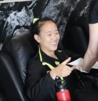 A smiling woman with shoulder-length black hair wearing a blue top standing among green plants in a bright indoor setting.