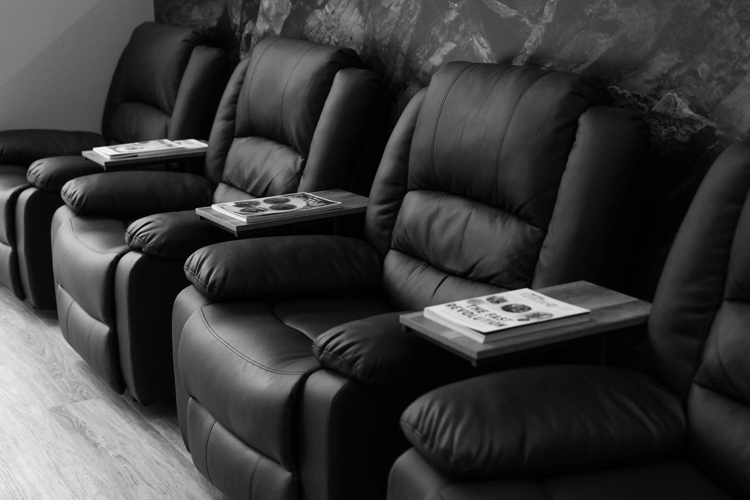 Row of black leather lounge chairs in a waiting area, each with a small wooden table holding magazines, with a scenic mountain mural on the wall behind.