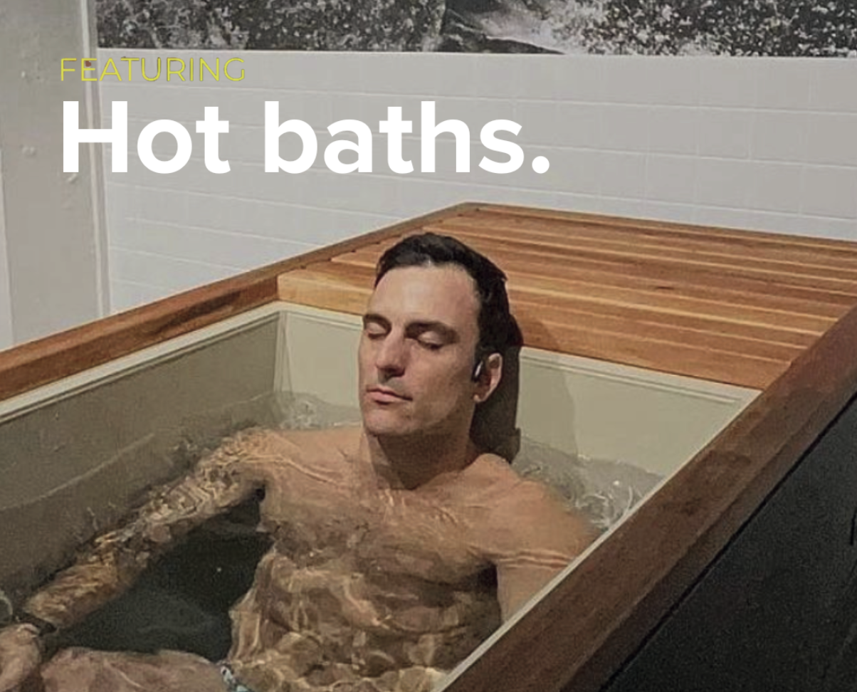 A man relaxing in a wooden hot bath with his eyes closed, surrounded by a white tiled wall, with text overlay that says "FEATURING Hot baths."