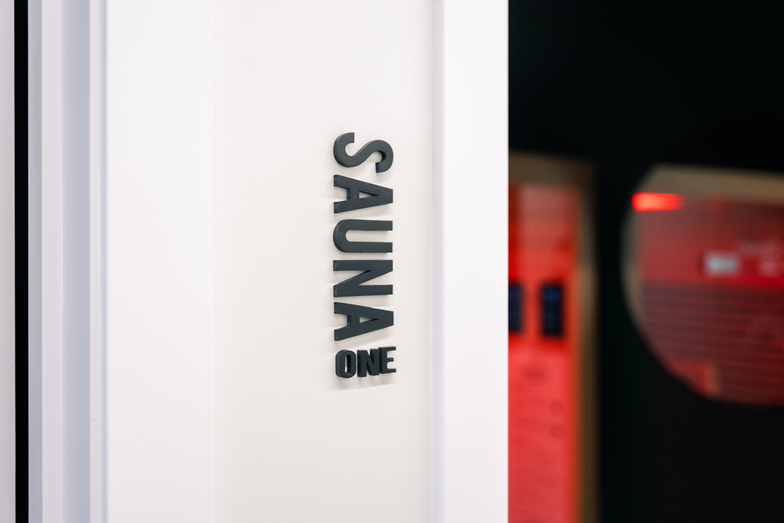 Sign that reads 'SAUNA ONE' on a white wall, with a blurred background including red and black colors.
