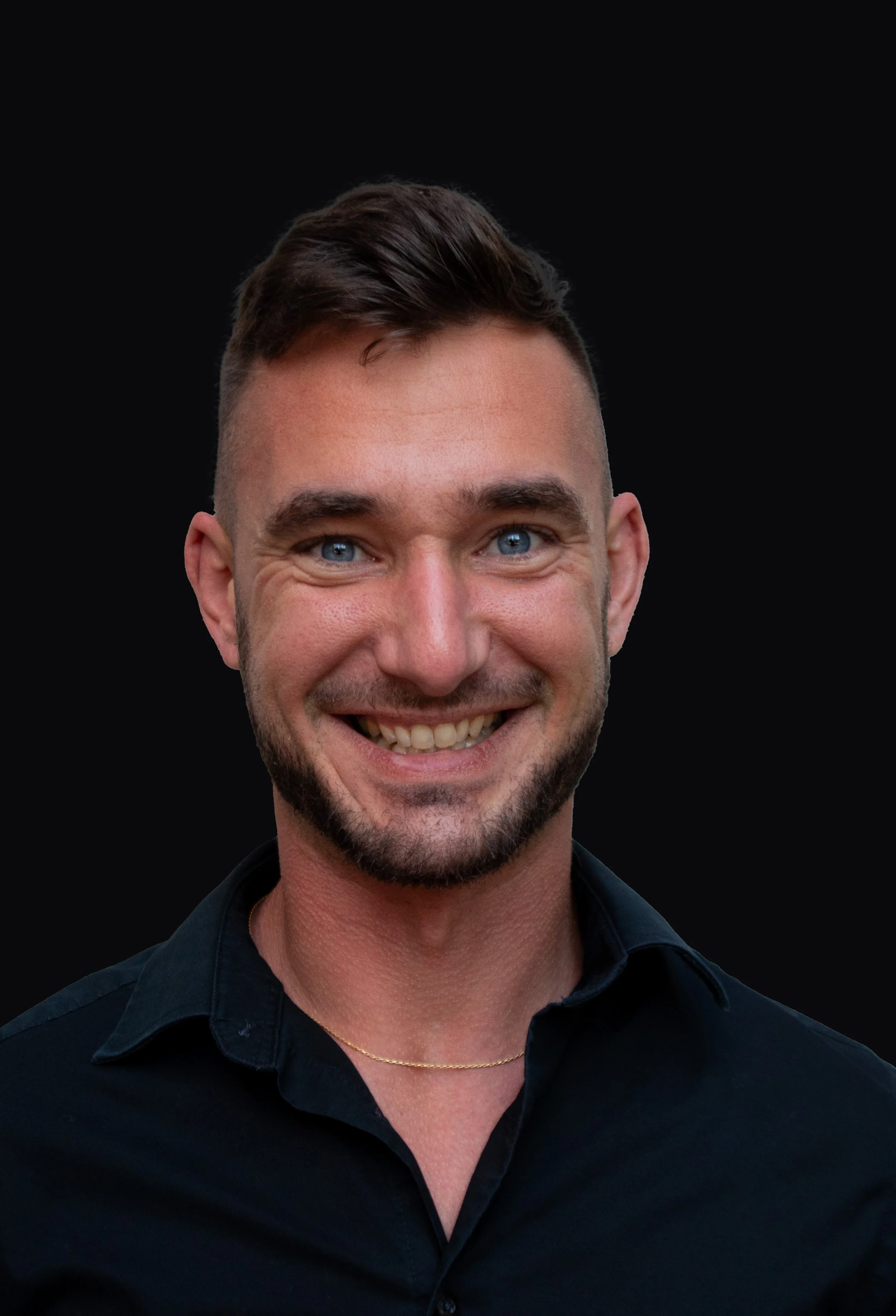 A smiling man with blue eyes, dark brown hair, and a beard, wearing a black shirt and a gold necklace, against a black background.
