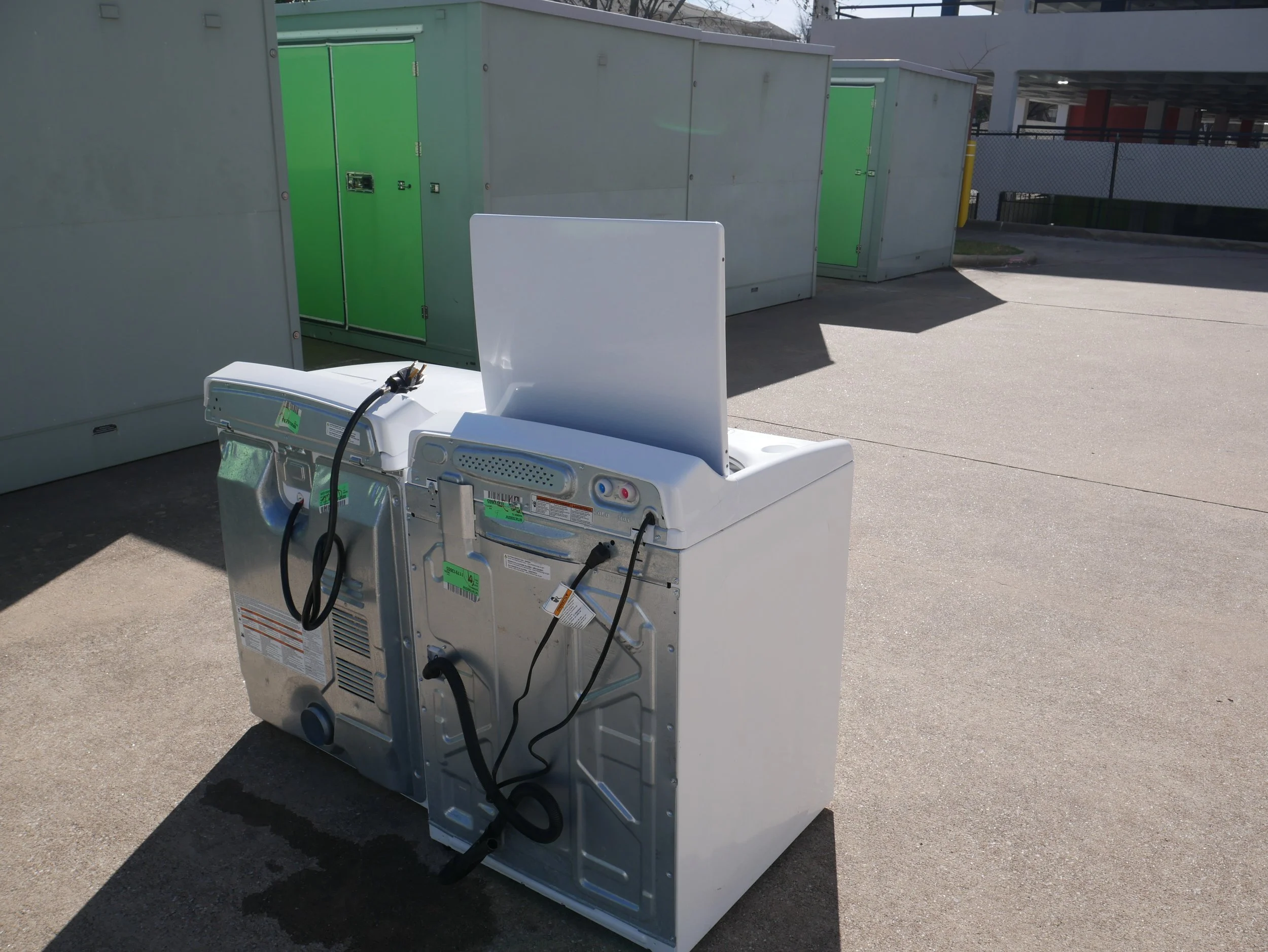 A commercial washer machine with its lid open, situated outdoors on pavement, with a white curtain or panel attached to the back, and green utility boxes or storage units in the background.