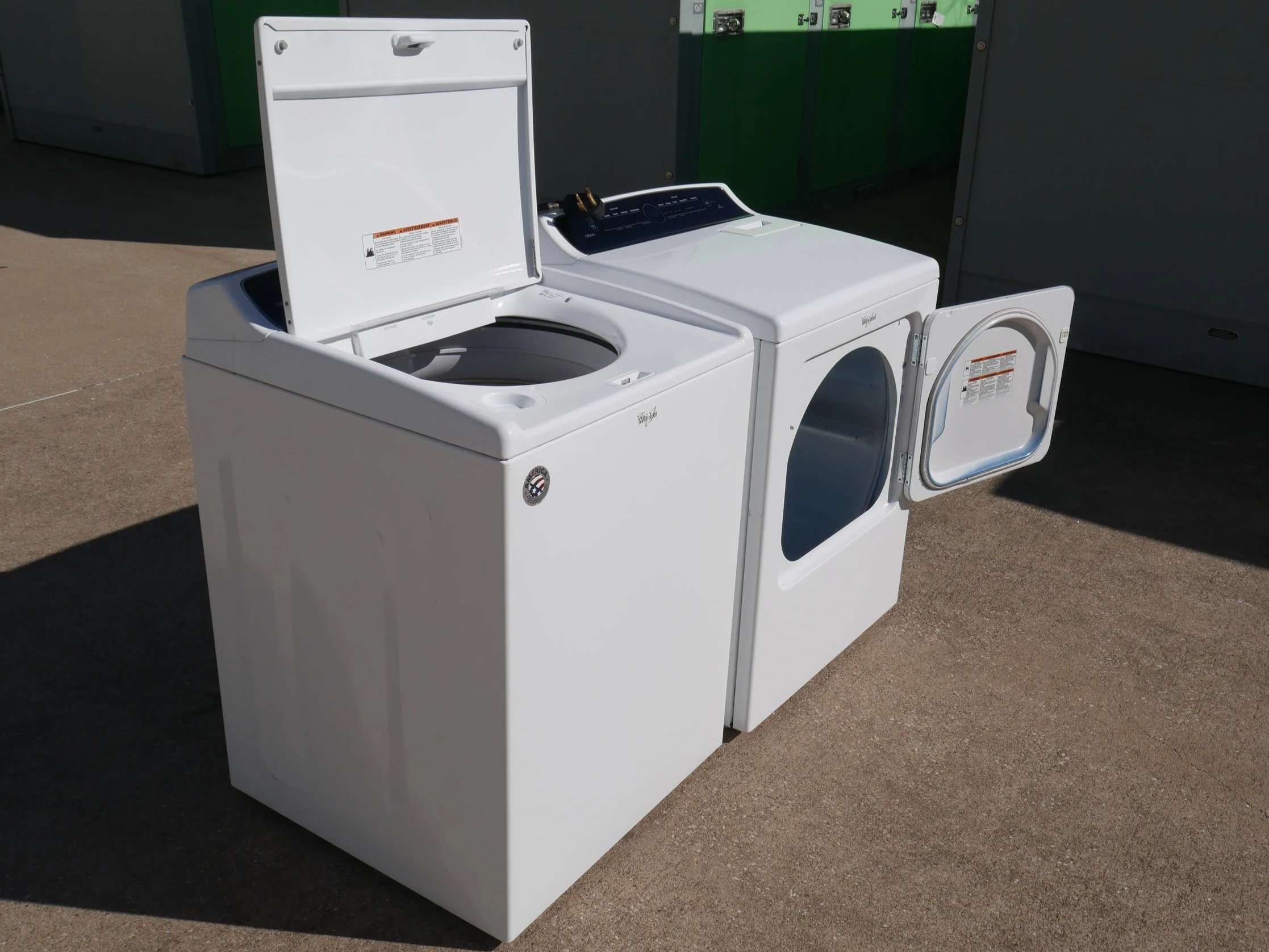 A white top-load washing machine and a white front-load dryer placed side by side outdoors on a concrete surface, with the dryer door open.