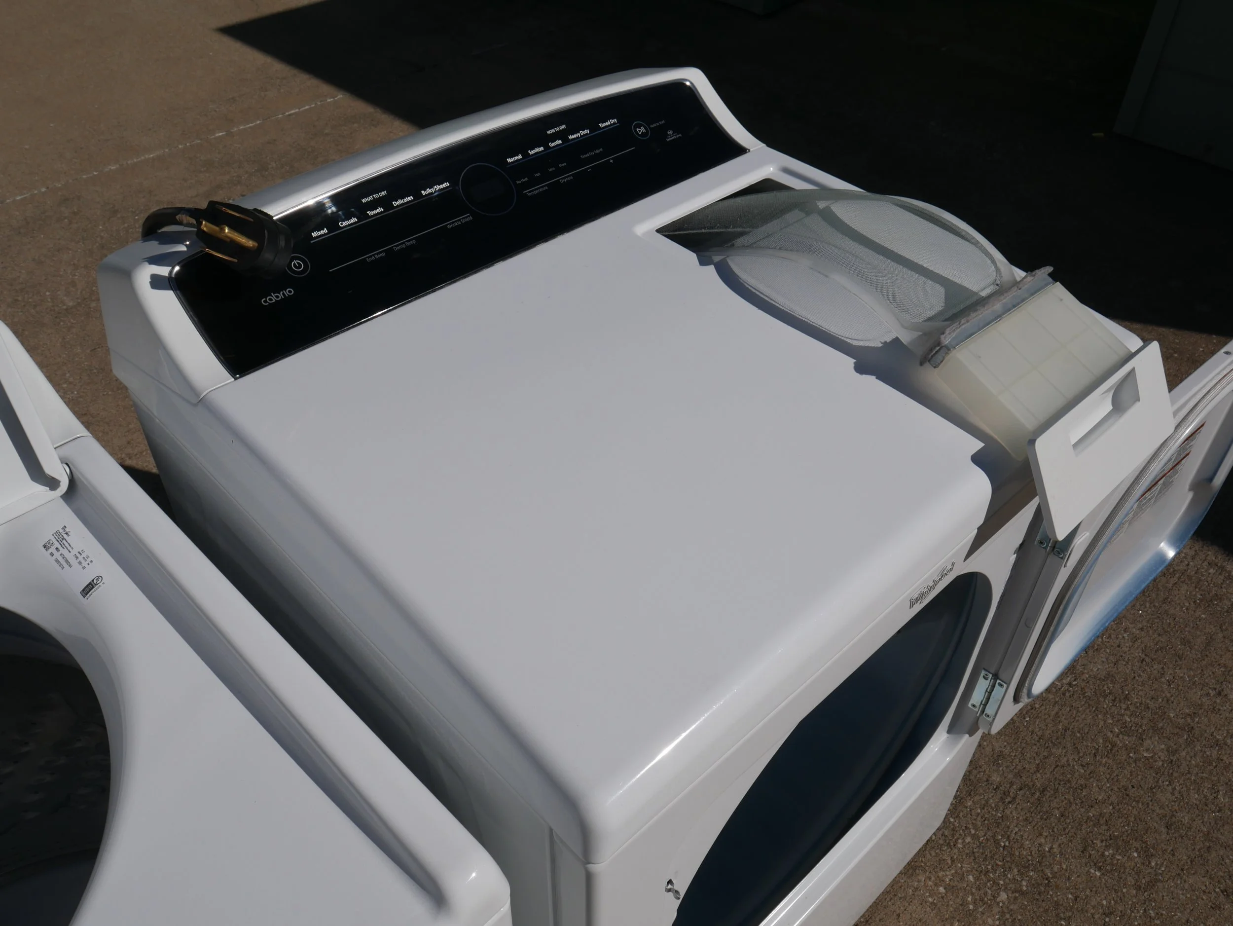A white top-loading washing machine with a control panel on top, located outdoors on a concrete surface, with a laundry lint collector attached to the top lid.