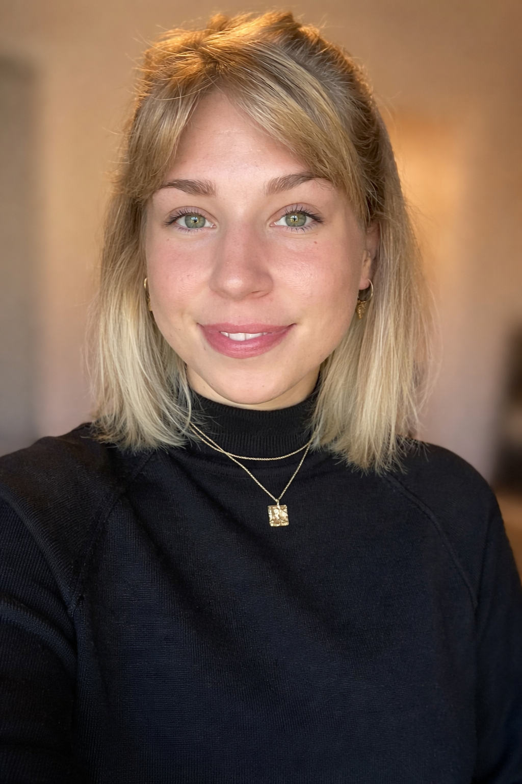 A woman with blonde hair and green eyes smiling at the camera, wearing a black top and gold jewelry.
