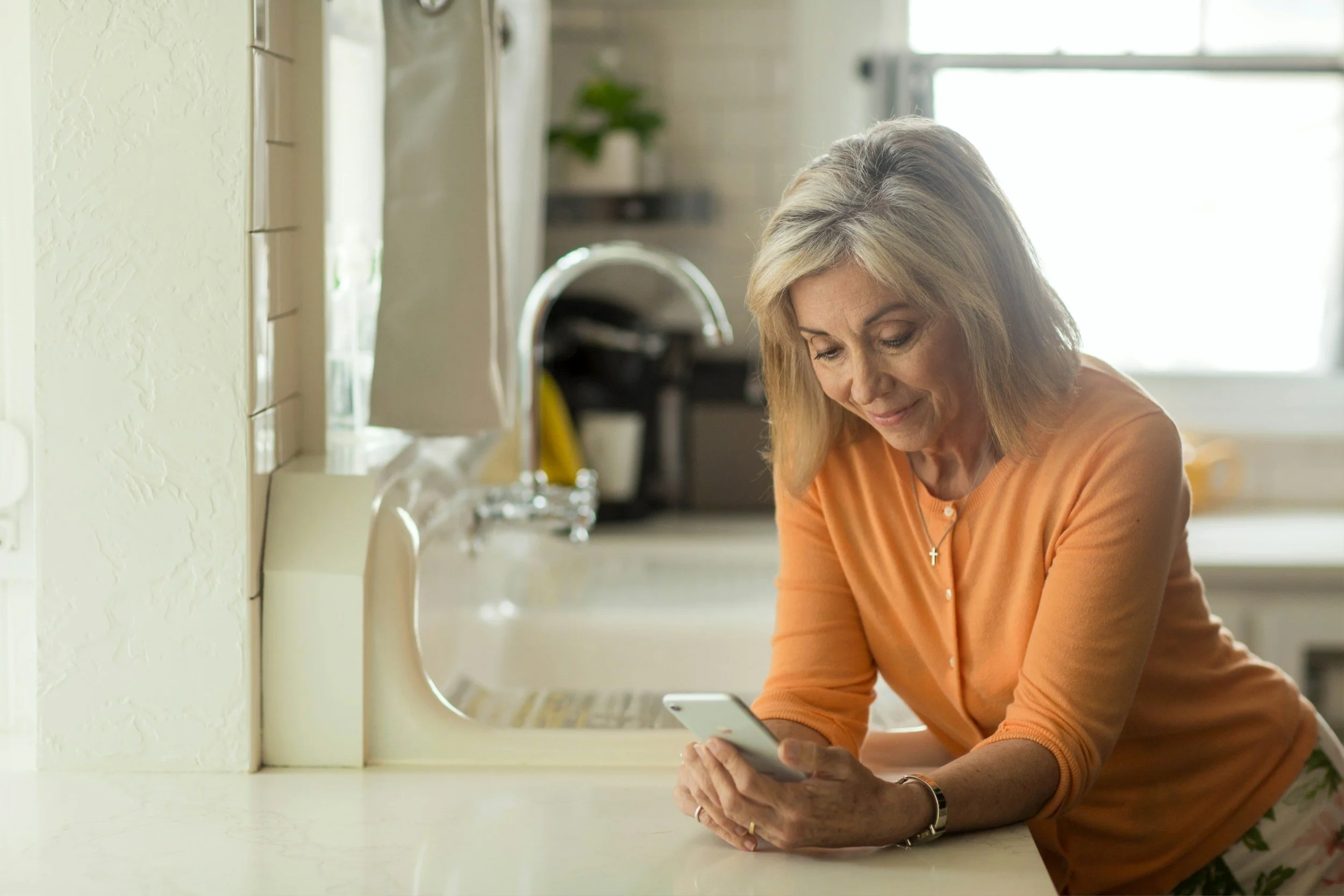 A woman with blonde hair wearing an orange top is leaning on a kitchen counter while looking at her smartphone.