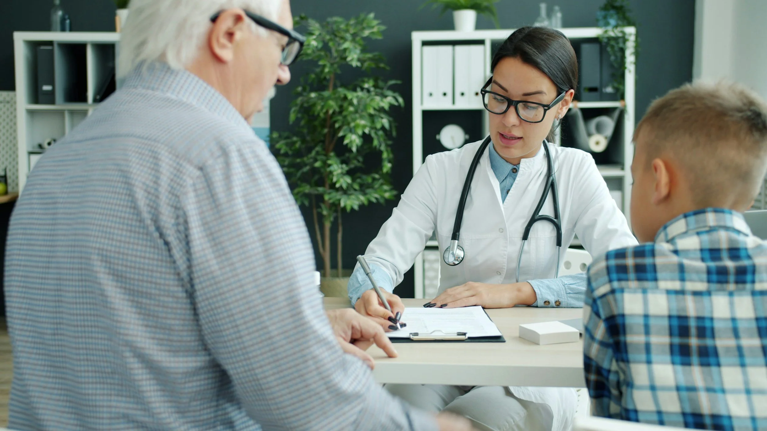 A young female doctor with glasses and a stethoscope around her neck speaking to an elderly man and a young boy in a medical office.