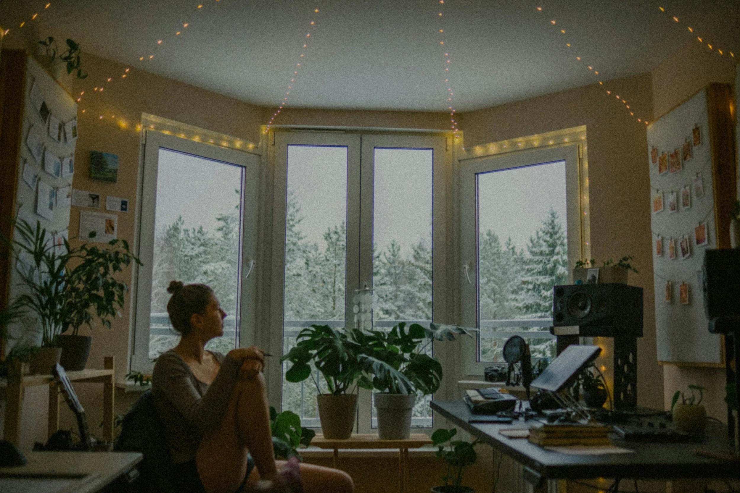 A woman sitting in a cozy, dimly-lit room with large windows, surrounded by plants, computer equipment, and string lights, with a snowy outdoor scene visible through the windows.