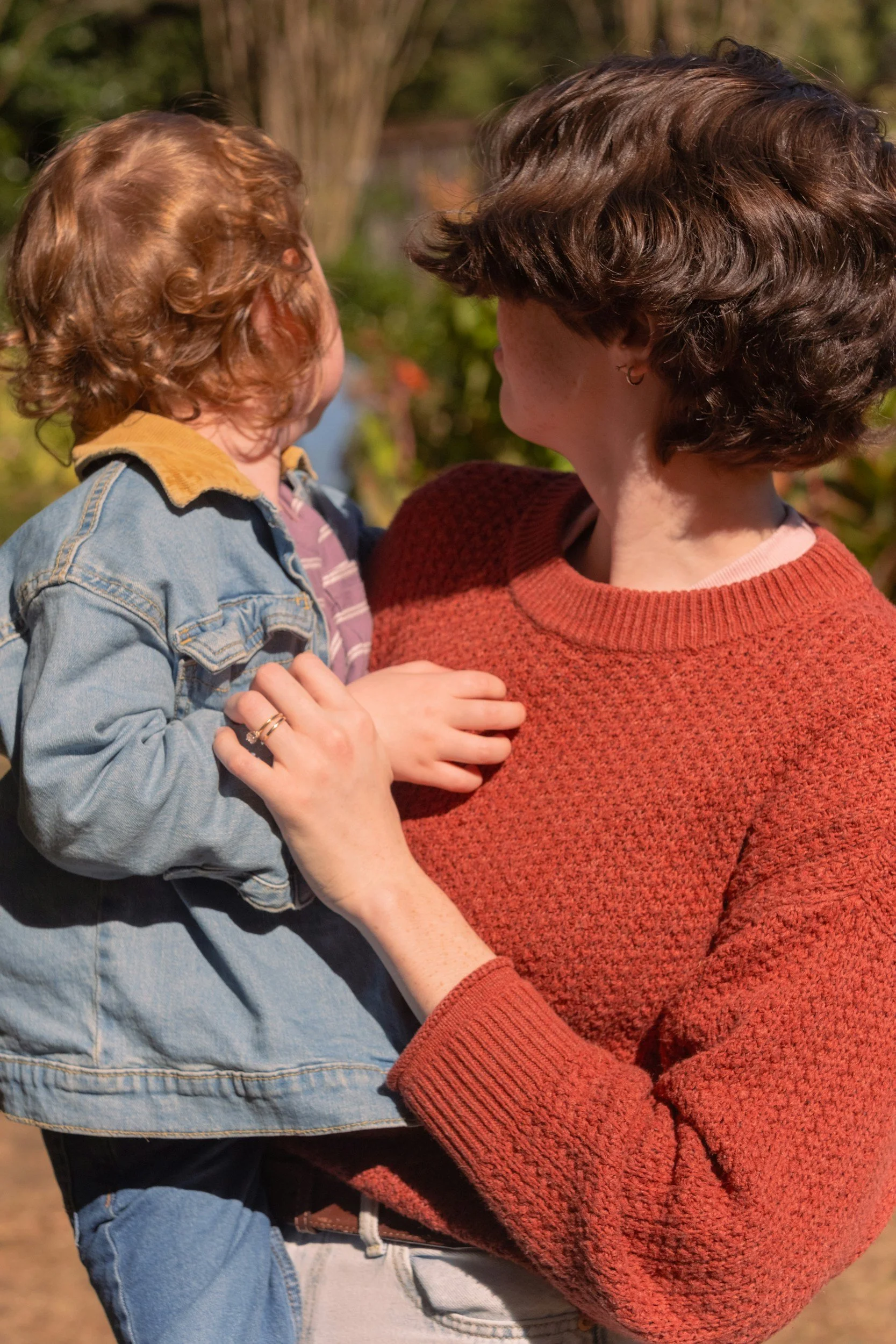A woman holding a young child outdoors on a sunny day, with trees in the background.