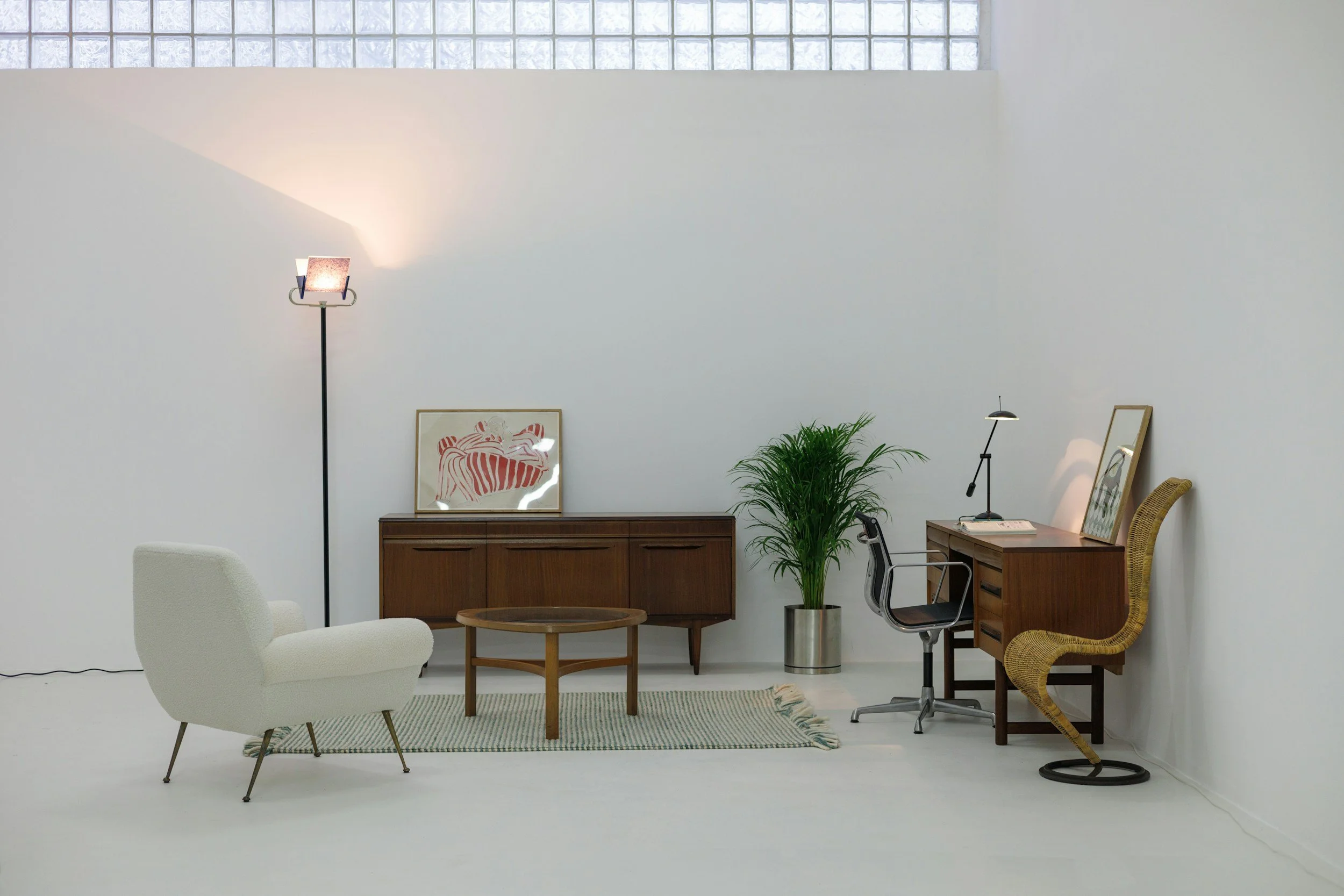 Minimalist living room with white walls, a white armchair, a wooden coffee table, a rug, a wooden sideboard with artwork, a tall floor lamp, a potted plant, and a desk with a chair and a small table lamp.