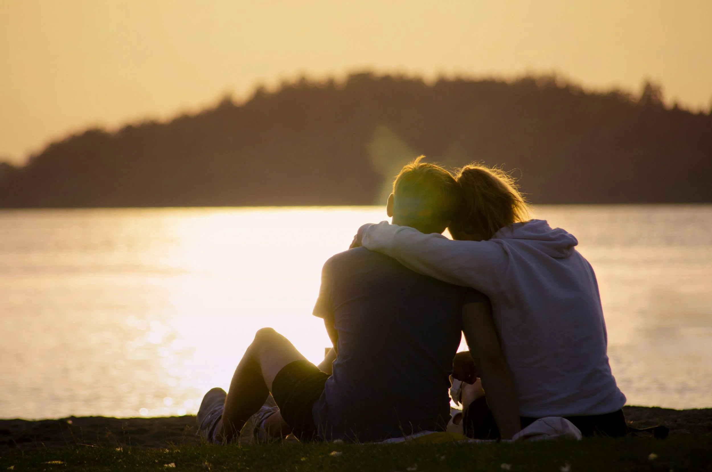 A couple sitting on a beach at sunset, embracing and watching the water and distant land.