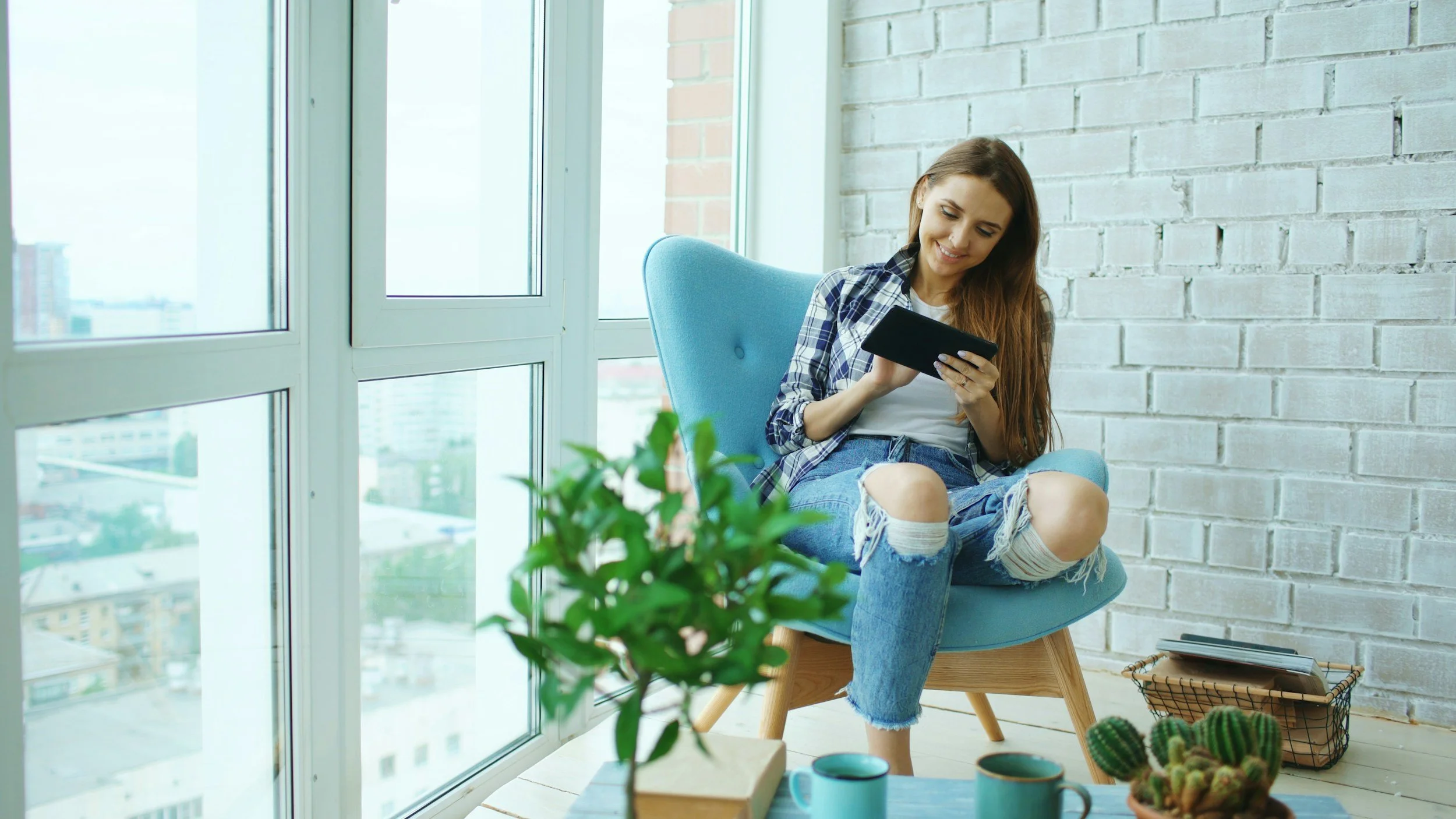 A young woman sitting in a blue armchair near a large window, smiling and looking at her phone. There are potted plants and cups on the table in front of her, and a basket with a phone inside on the floor nearby.