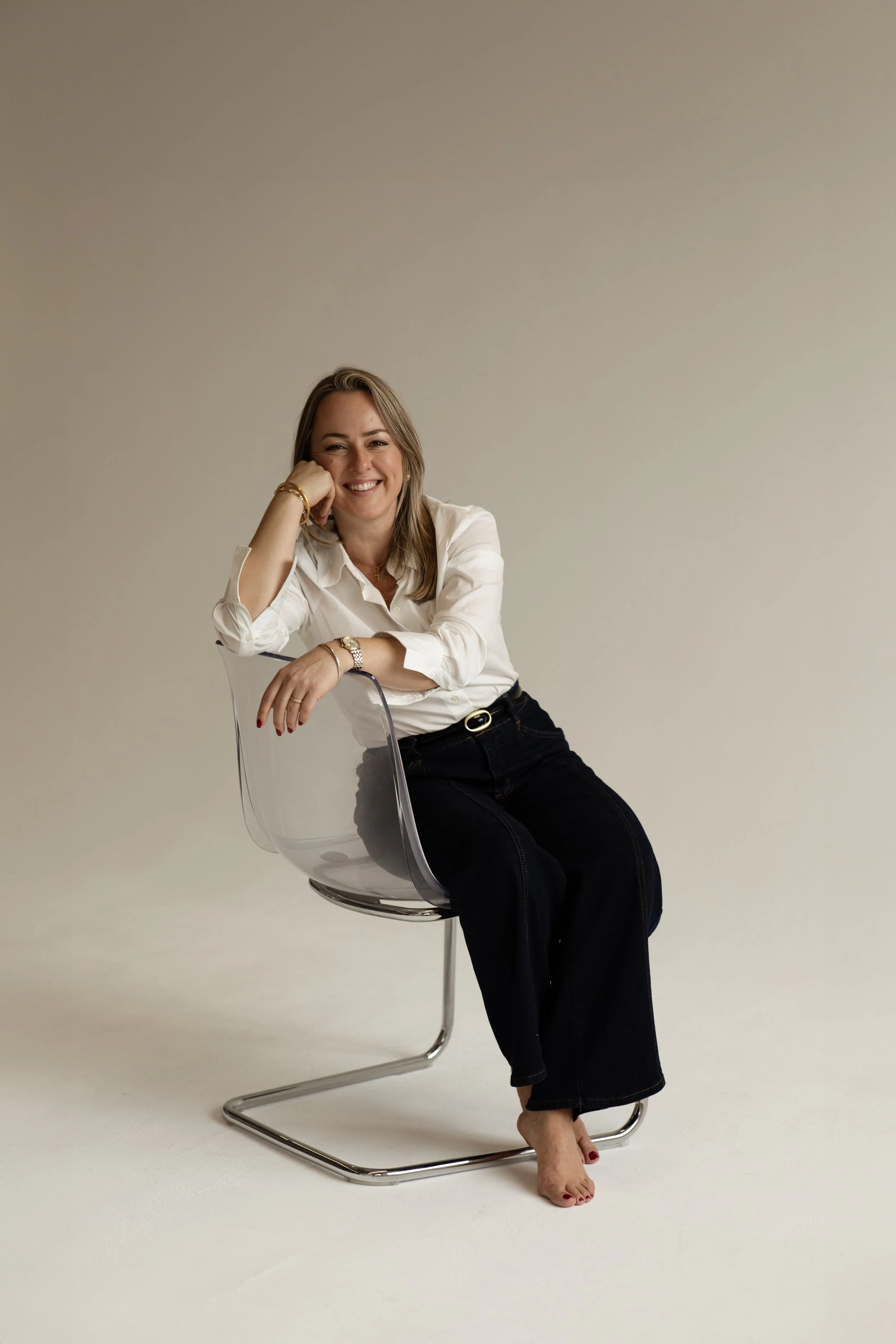 A woman sitting on a transparent chair, smiling with her head resting on her hand, wearing a white blouse and black pants, with jewelry and painted toenails, against a plain background.