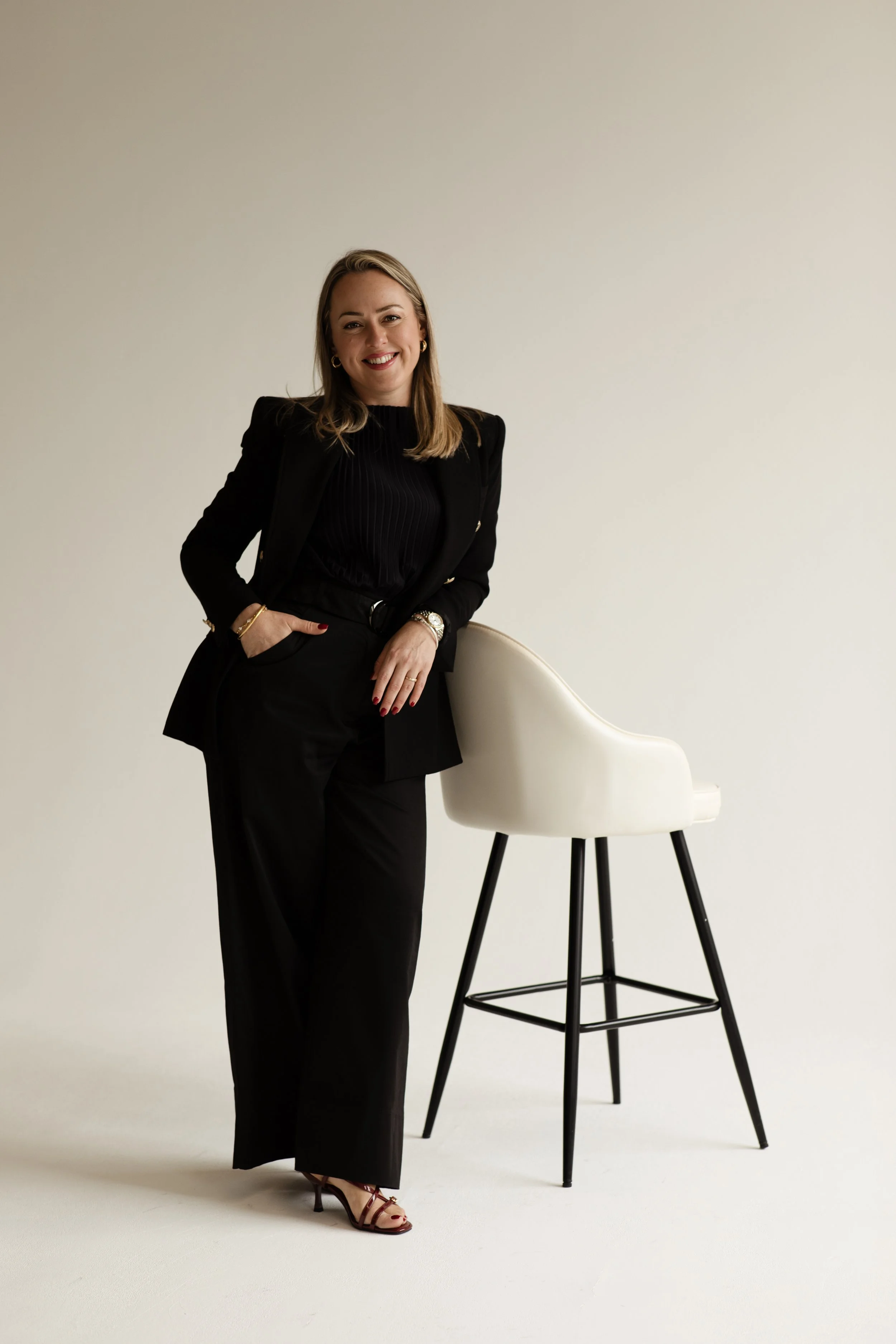 A woman in a black suit with a black top, standing next to a white chair with black legs in front of a plain light-colored background, smiling at the camera.