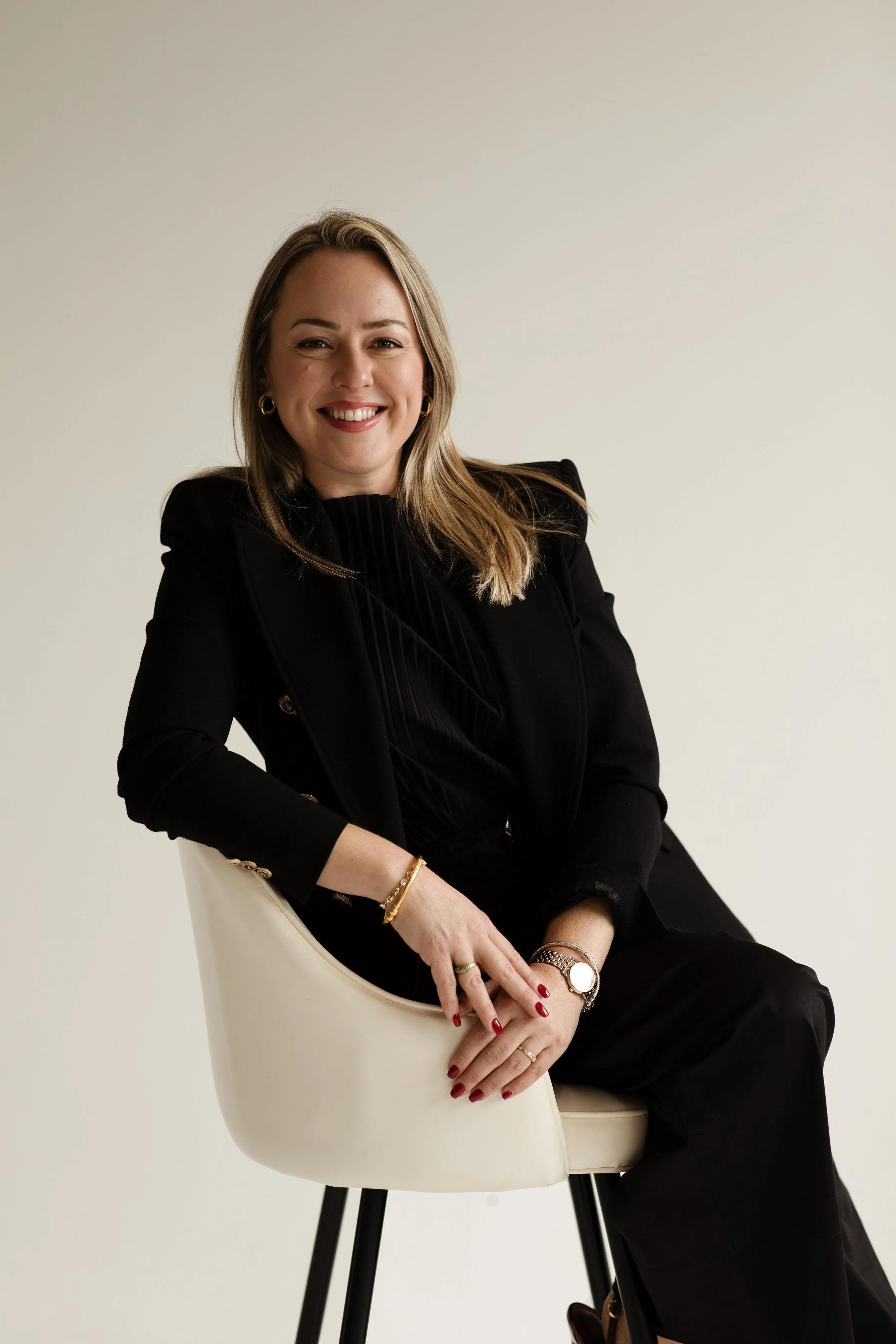 A woman with blonde hair, wearing a black blazer and pants, sitting on a modern white chair with black legs, smiling at the camera against a plain light background.
