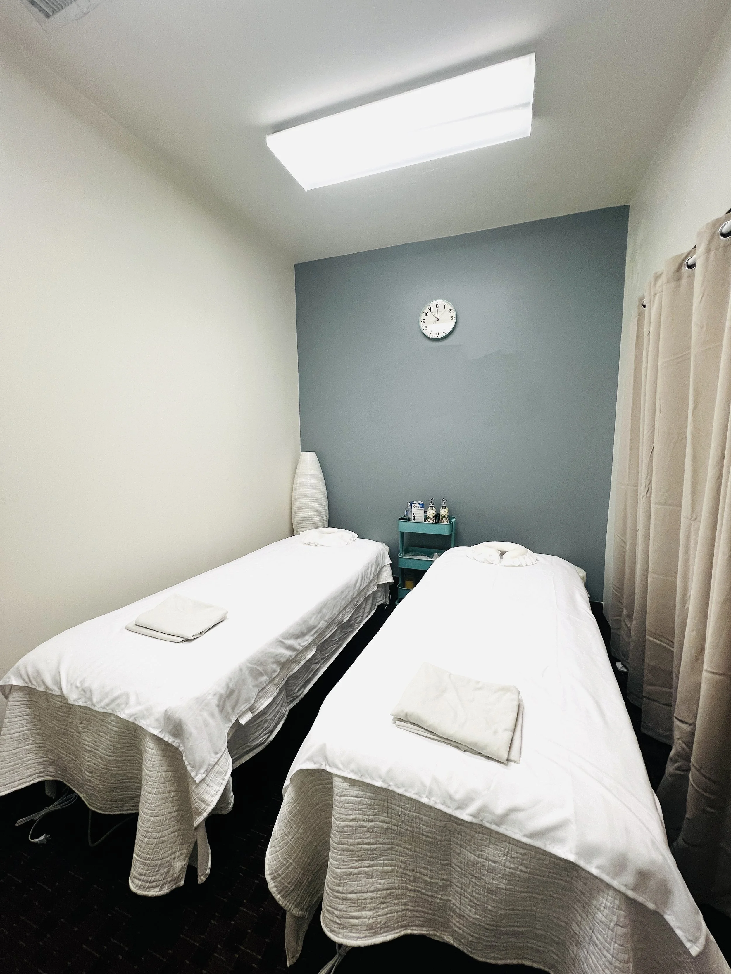 A hotel room with a bed, a tall white vase, a wall clock, a framed object, a nightstand with a lamp, a small plant, and various bottles and items on the nightstand. The room has a beige wall and dark carpeting.